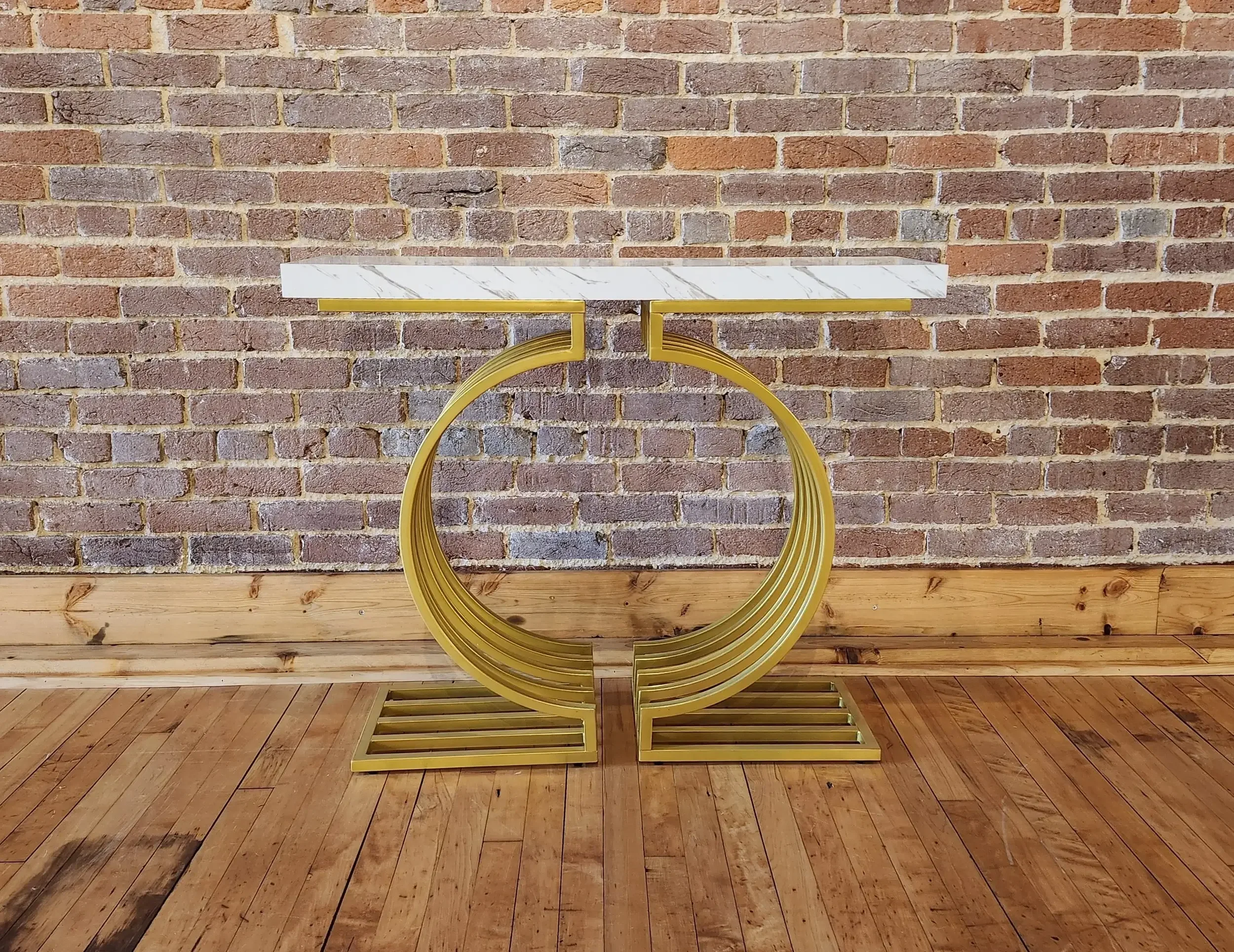 A gold-colored modern side table with a white marble top against a brick wall and wooden floor.