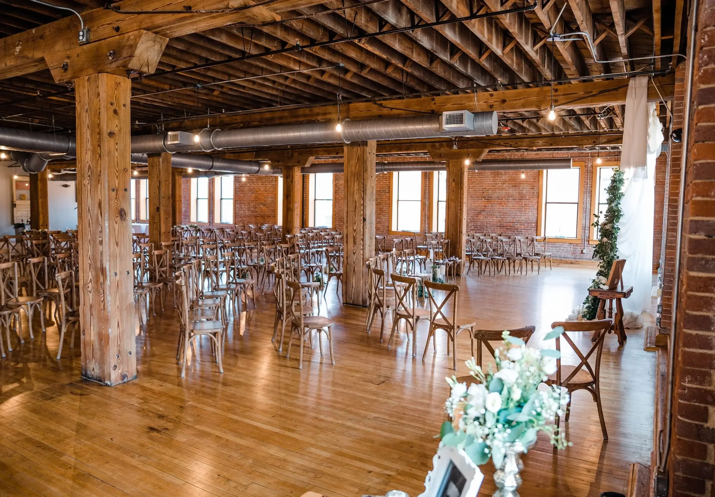 An empty event space with wooden floors and exposed brick walls, set up with multiple rows of wooden chairs, decorated for a wedding or special occasion, with floral arrangements and natural light coming through large windows.