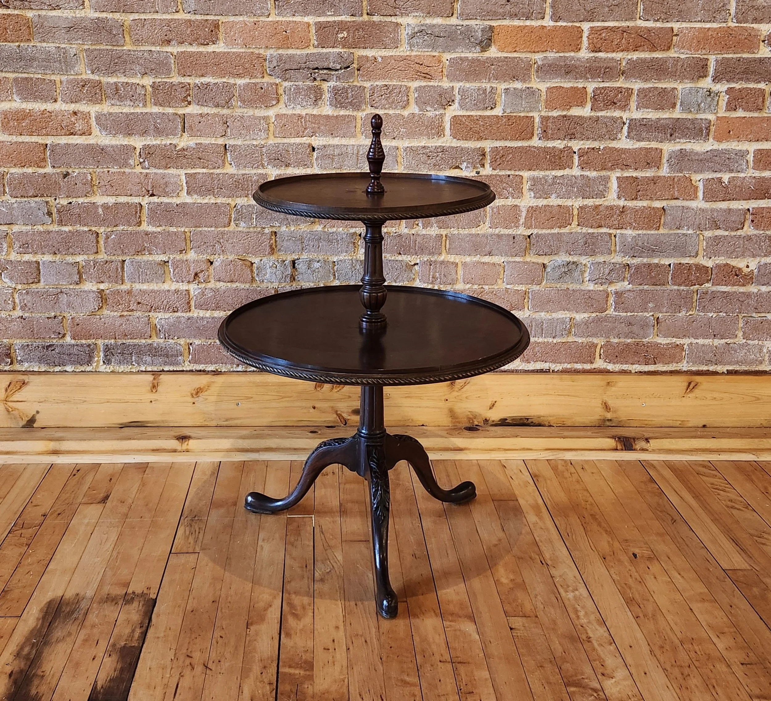 A three-tiered dark wood side table with curved legs, standing on a wooden floor against a brick wall background.