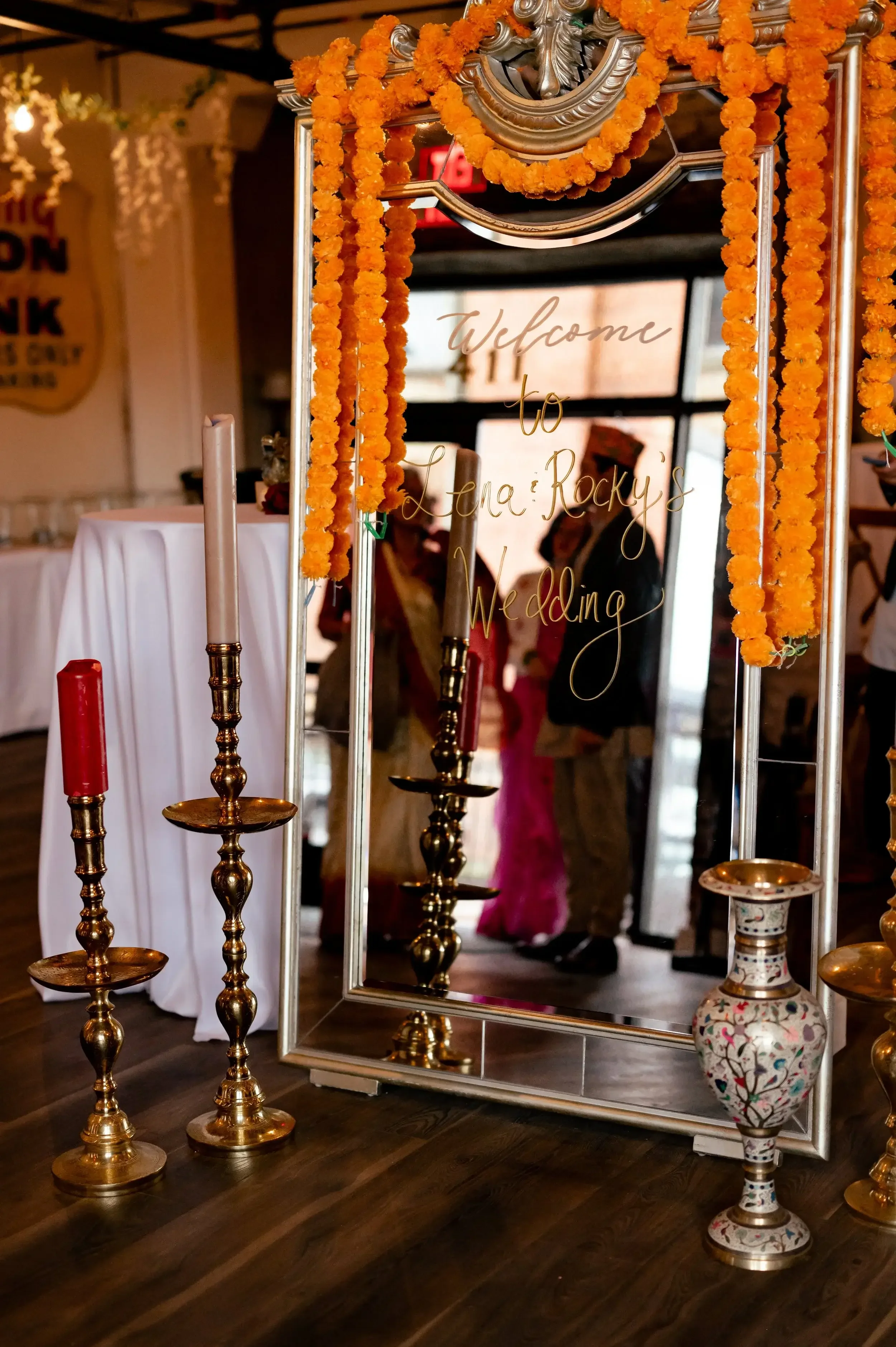 A mirror decorated with orange marigold flowers and a handwritten sign that reads "Welcome to Lena Rocky's Wedding." Several tall candle holders with red and pink candles are placed in front of the mirror, and a floral-patterned vase is seen on the r