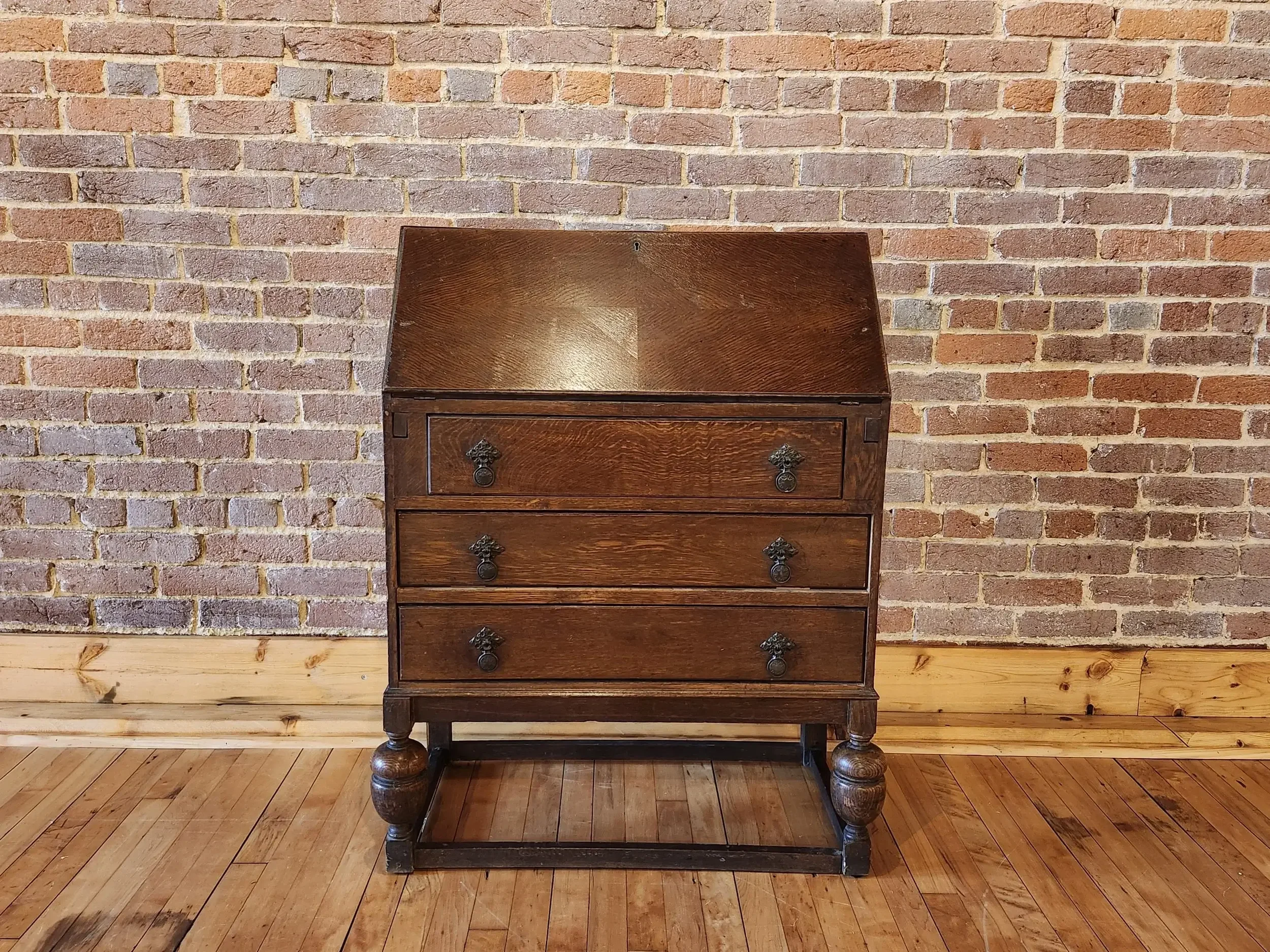 A vintage wooden writing desk with a slanted top, four drawers with ornate metal handles, and decorated legs, placed against a brick wall and wooden floor.