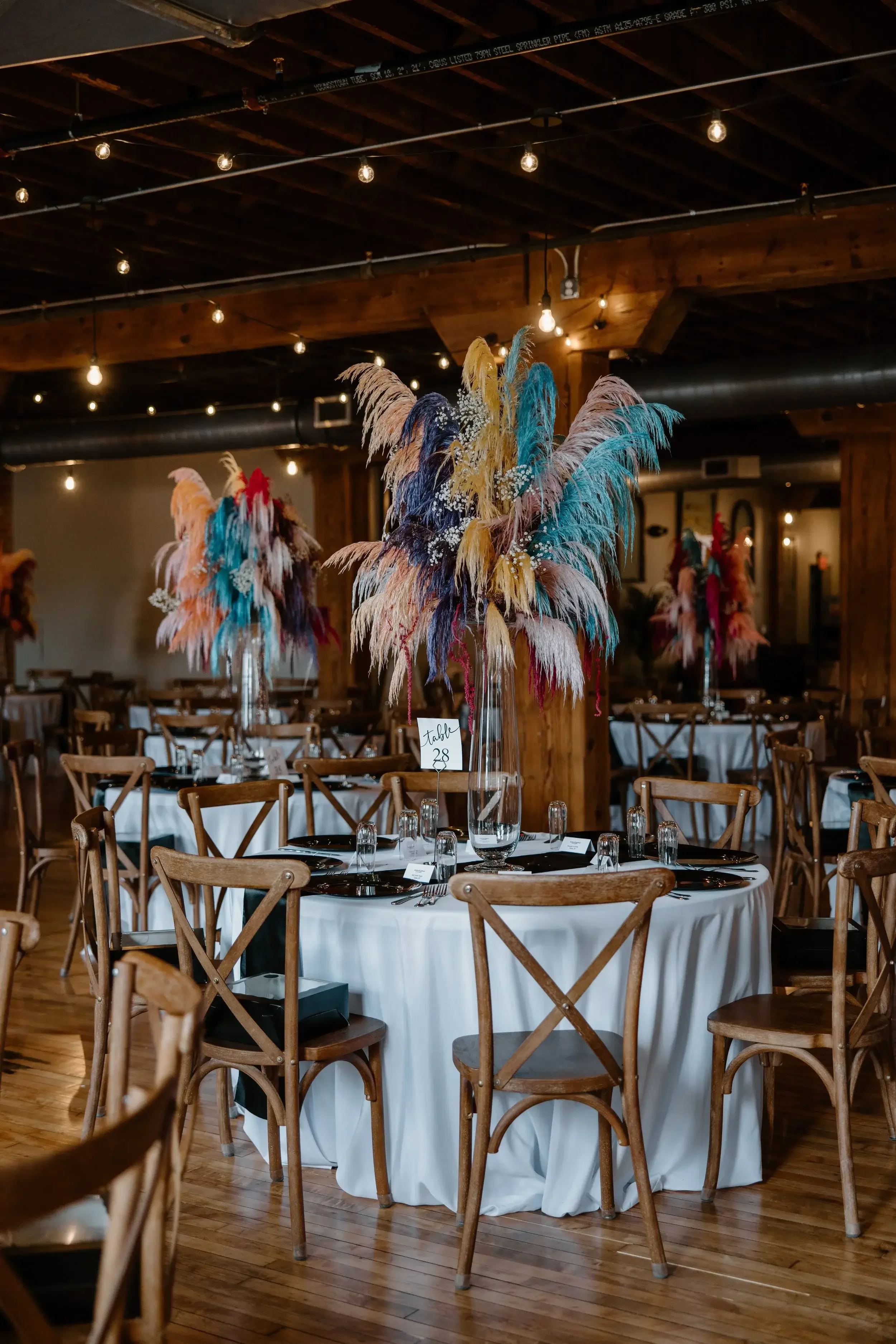 Wedding reception table decorated with a tall clear vase filled with colorful feather arrangements, surrounded by wooden chairs and set with black plates and silverware, inside a rustic wooden venue with string lights.
