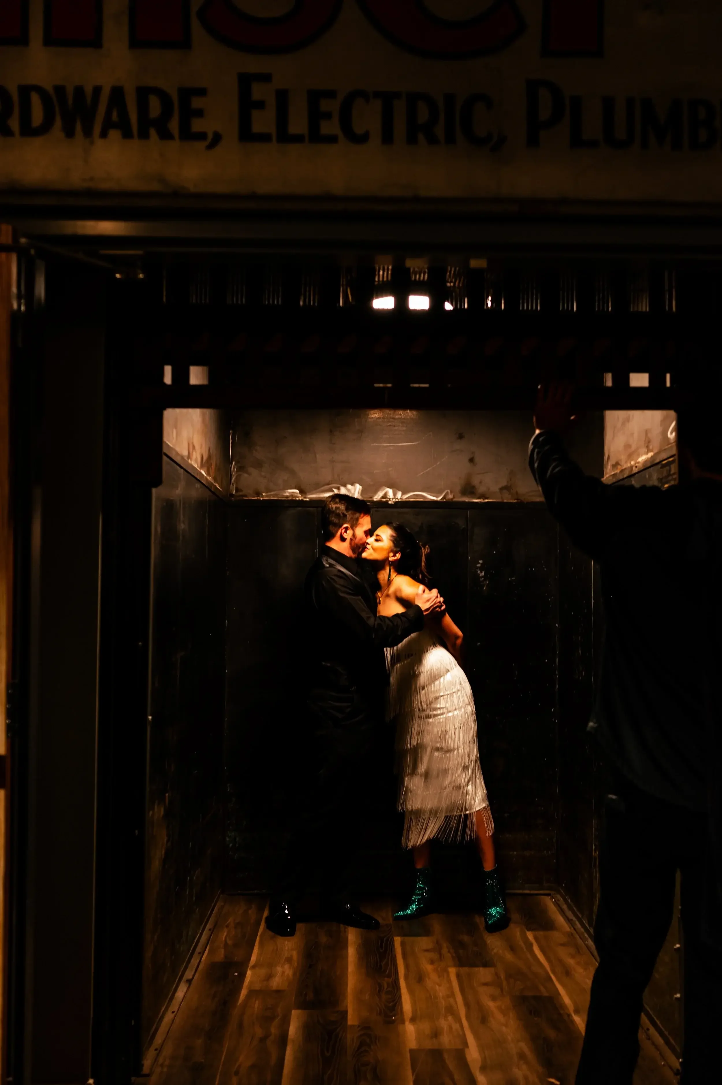 A couple is kissing in a dimly lit elevator, with the woman wearing a white dress and shiny boots, and the man in dark clothing. A person on the right is taking the photo.