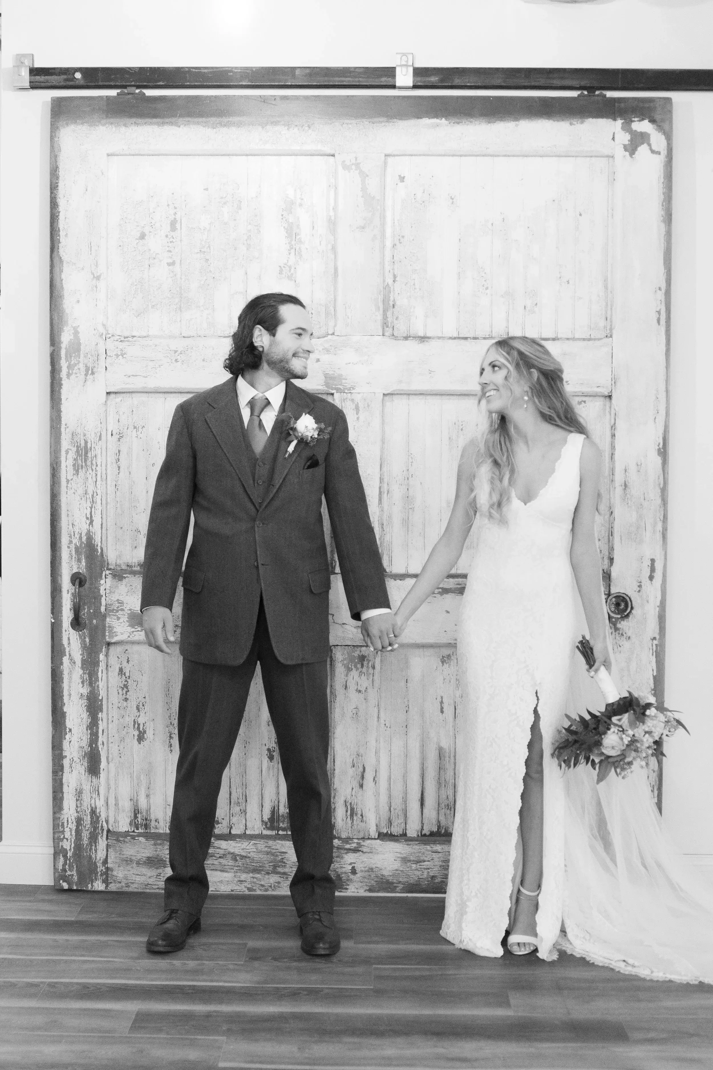 A black and white photo of a bride and groom holding hands and smiling at each other, standing in front of a rustic, weathered wooden door with sliding track.