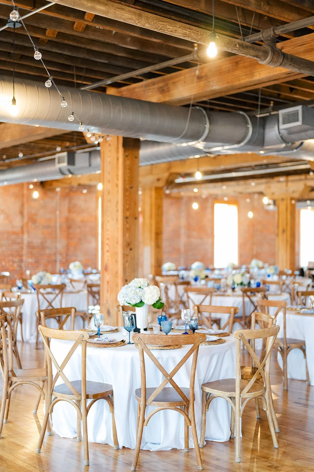 A decorated banquet hall with round tables draped in white tablecloths, each set with glassware, plates, and floral centerpieces, featuring wood chairs, exposed brick walls, wooden beams, ductwork, and hanging light bulbs.