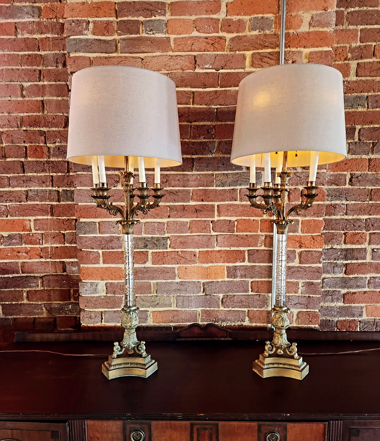 Two vintage brass table lamps with white fabric shades placed on a dark wooden surface against a brick wall.