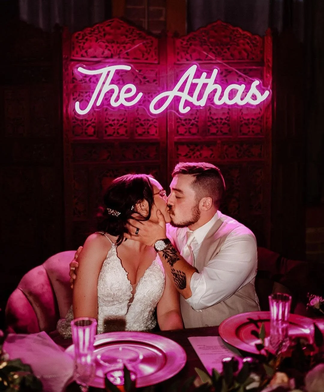 A couple sharing a kiss at a wedding reception, with a neon sign that reads 'The Athas' in the background.