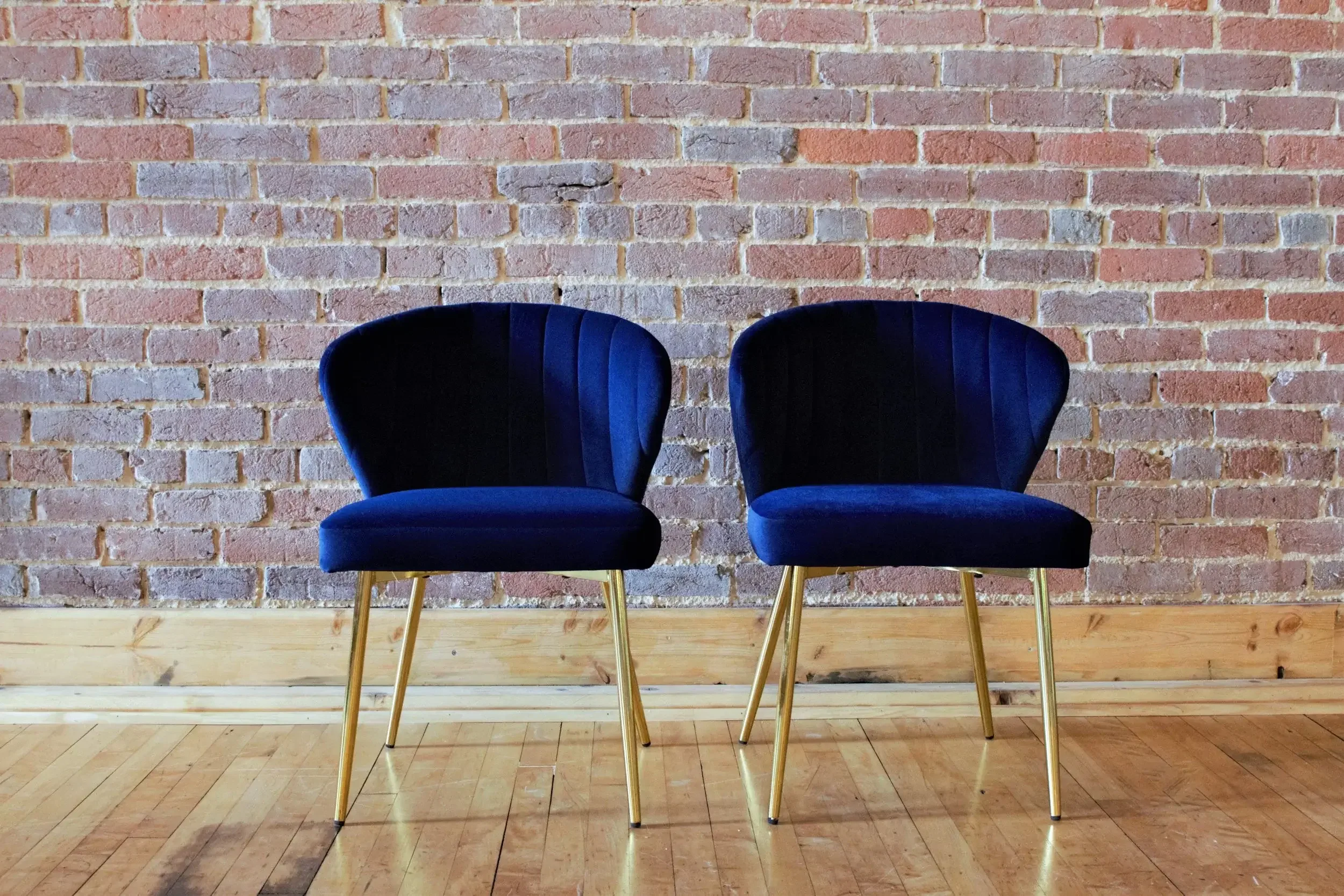 Two blue velvet chairs with gold legs placed against a red brick wall on a wooden floor.