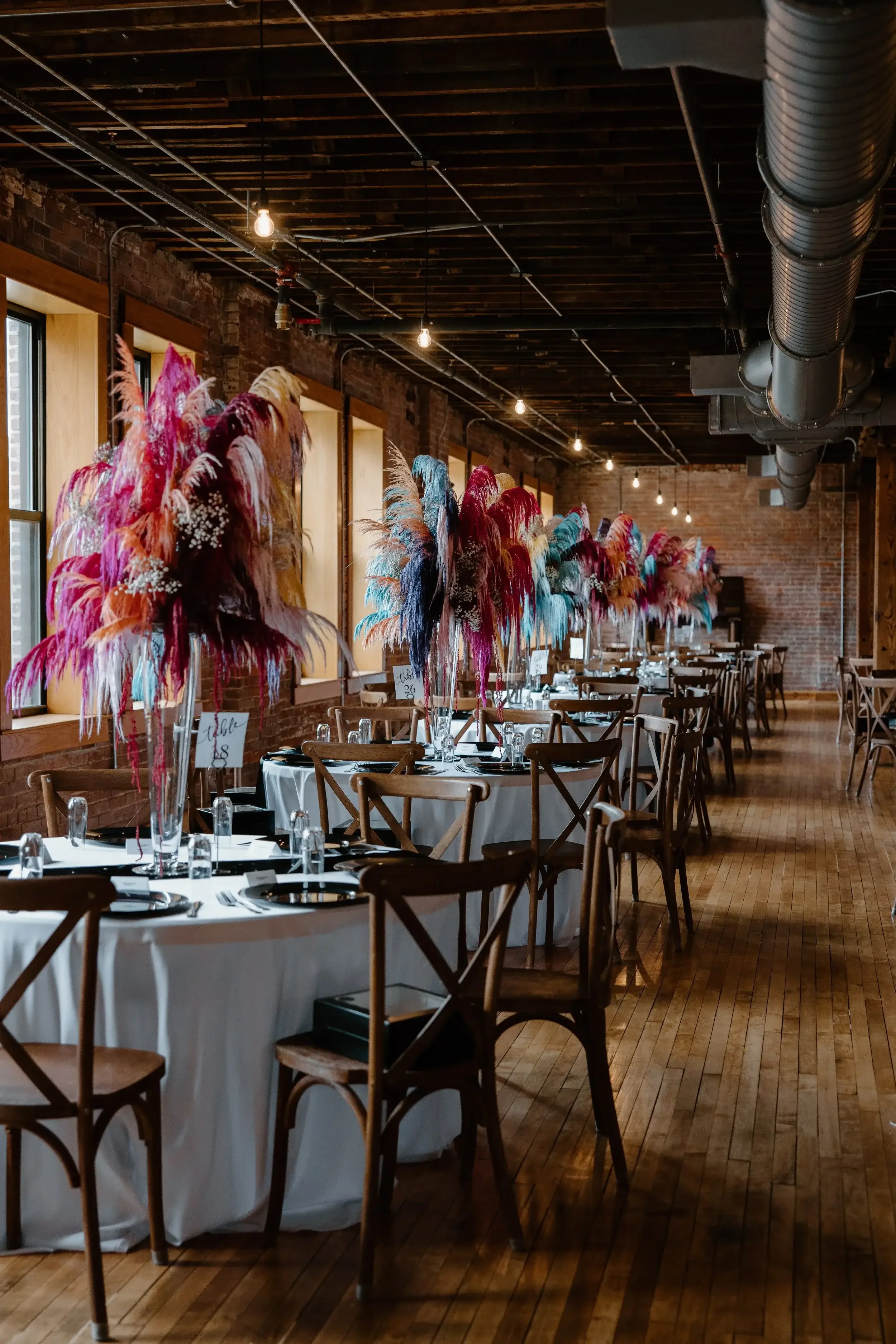 Wedding reception tables decorated with colorful feather centerpieces, set with black plates and glasses, in a rustic brick-walled venue with wooden floors and large windows.