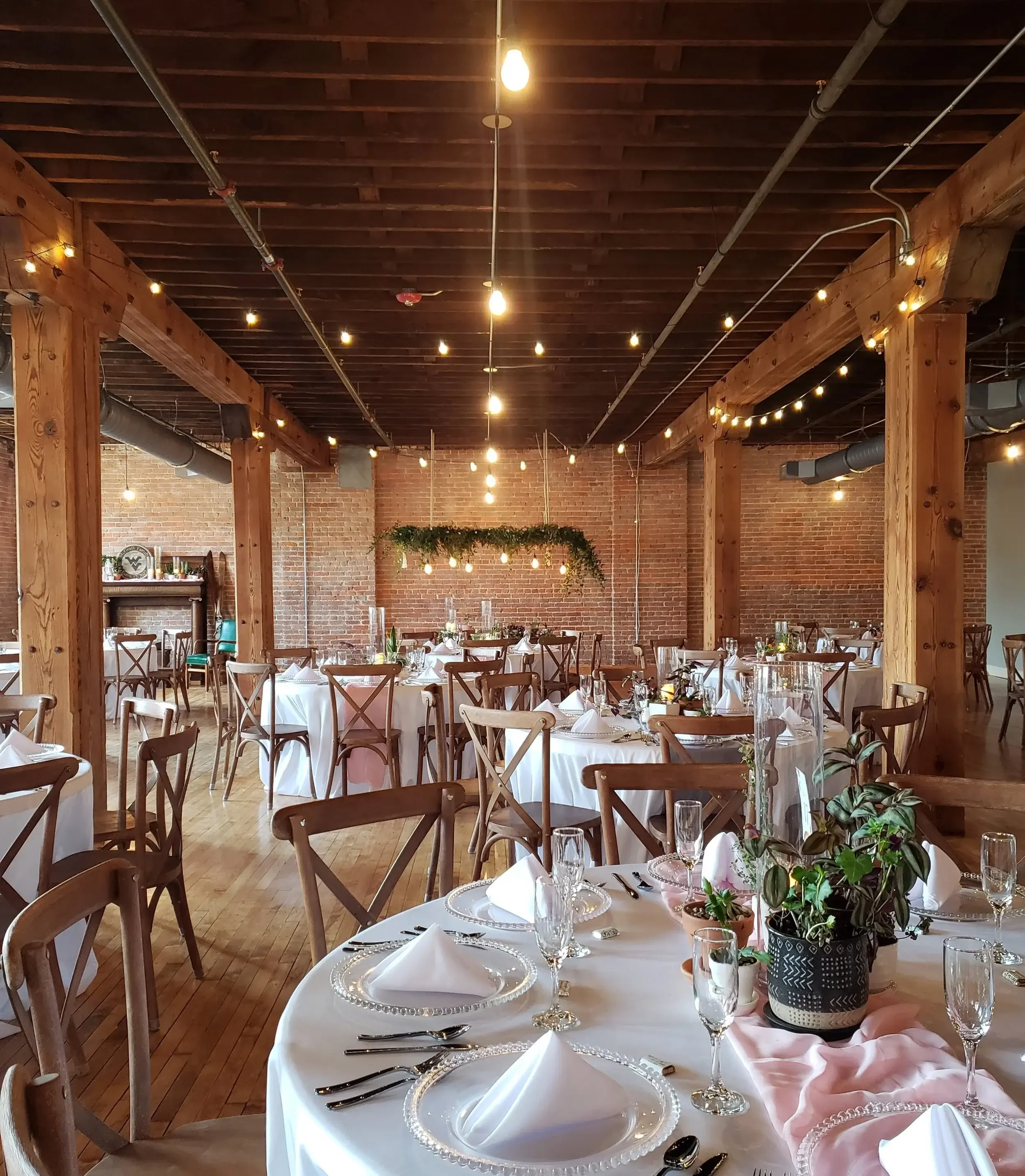Elegant event space with round tables covered in white tablecloths, set with folded white napkins, glassware, and silverware. There are potted plants on each table and decorative string lights hanging from the wooden beams of the ceiling. The room fe