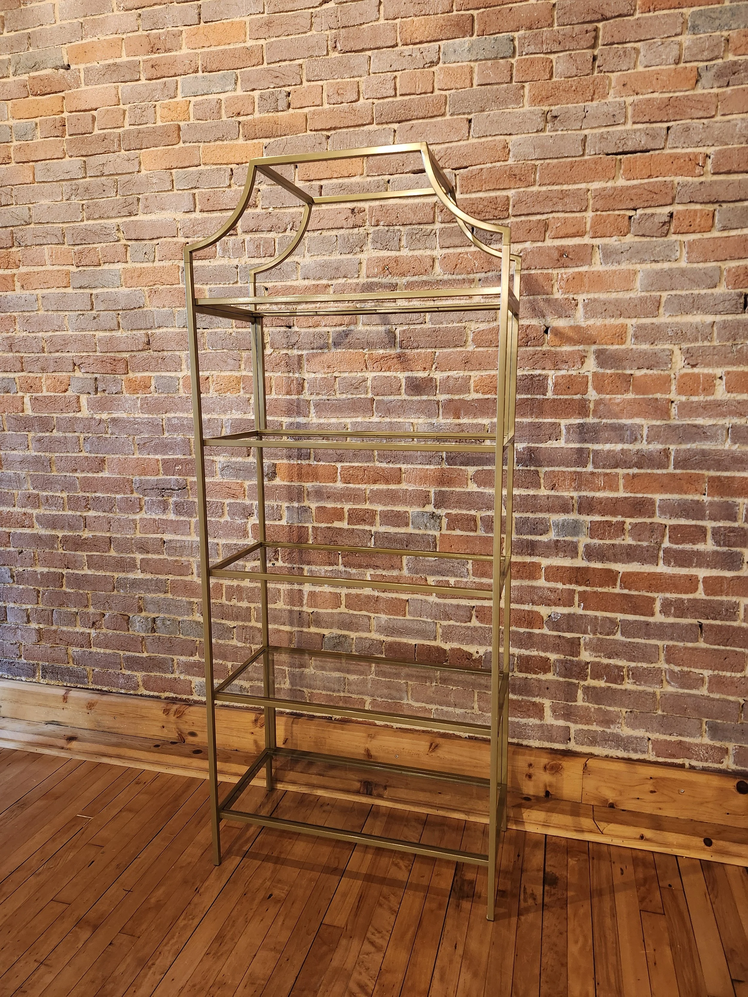 A tall, gold metal open shelving unit with five shelves, positioned against a brick wall and hardwood floor.