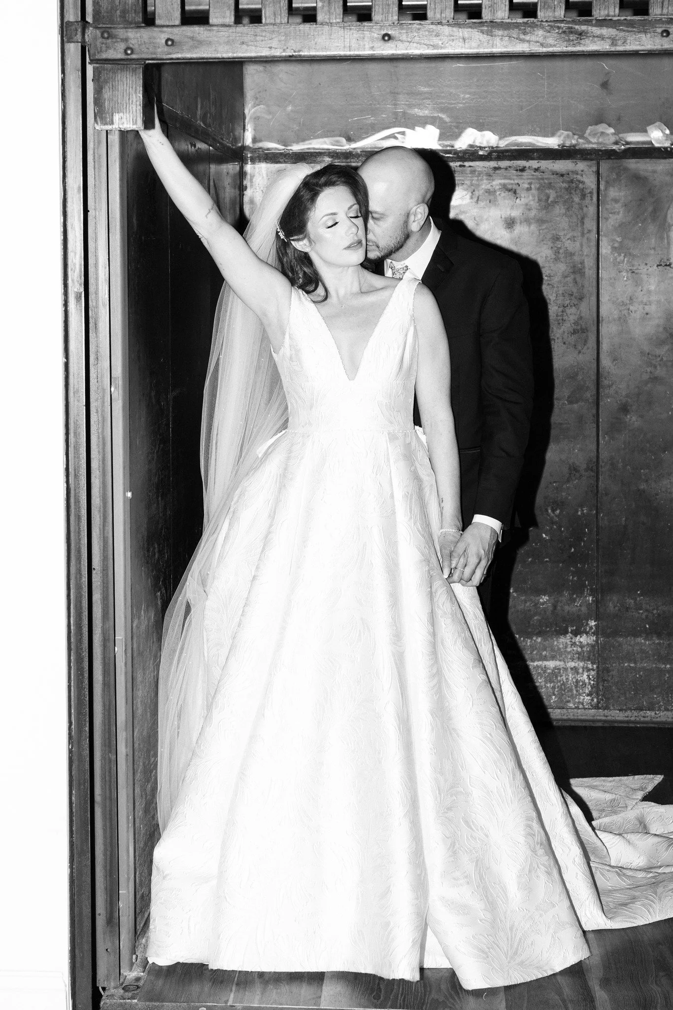 A bride and groom in wedding attire in a dark, industrial setting. The bride in a white gown with a deep V neckline and veil, her eyes closed, leaning against the groom. The groom in a black tuxedo, bald with a beard, standing close behind her, kissi