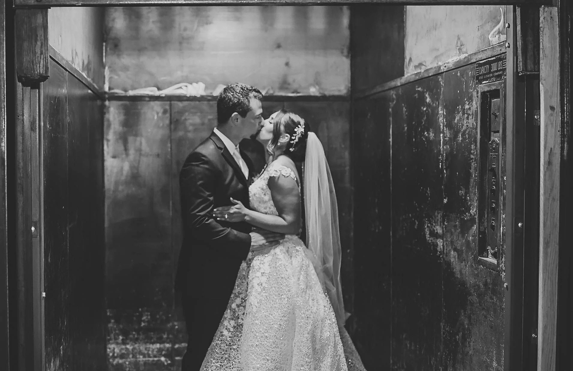 Black and white photo of a newlywed couple sharing a kiss in an industrial elevator with rustic walls.