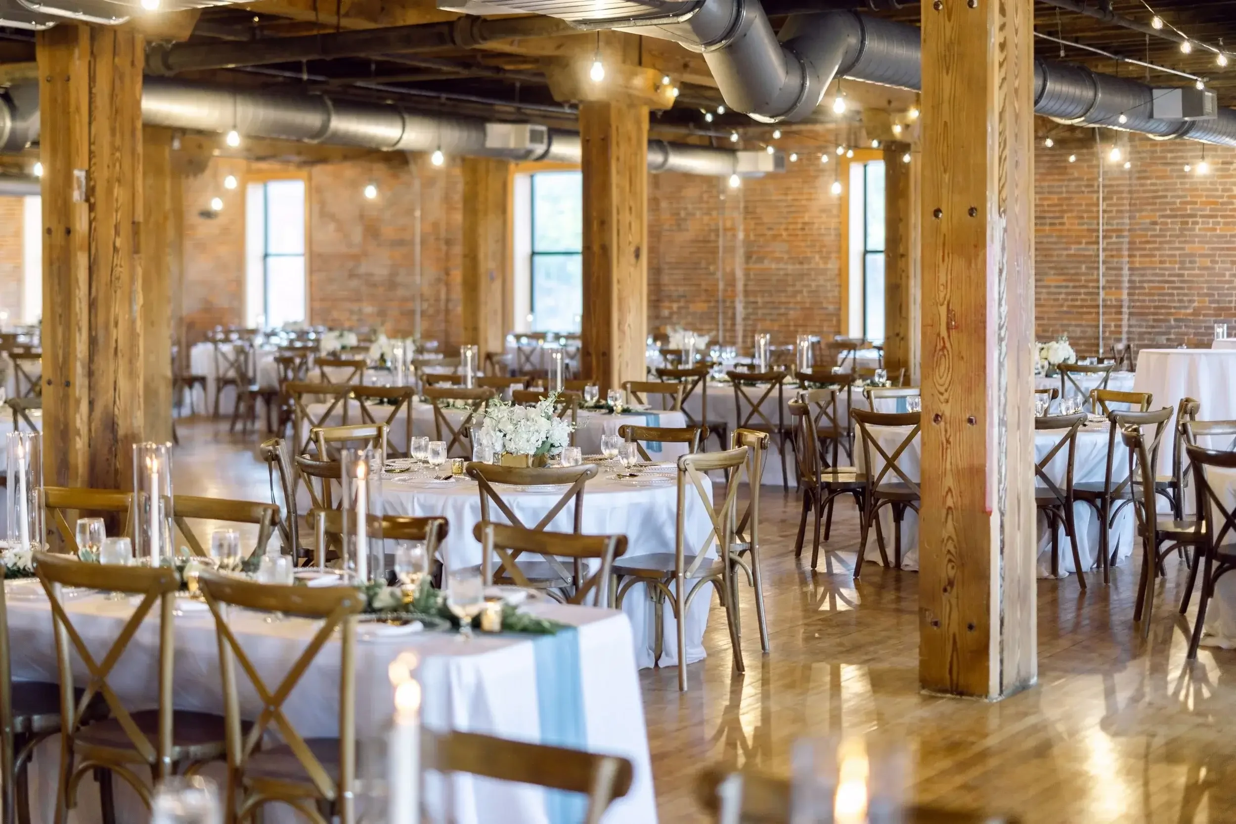 Interior of a rustic event space with wooden beams, brick walls, and large windows, set up for a formal gathering with round tables covered in white tablecloths, decorated with floral centerpieces and candles, and surrounded by wooden chairs.