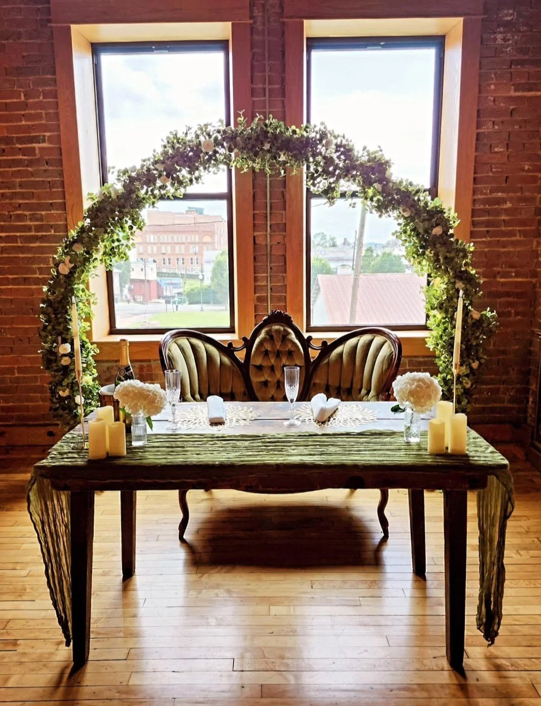 A decorated table set for a wedding or special occasion in a rustic room with brick walls and large windows. The table has floral arrangements, candles, champagne glasses, and a lace runner. A vintage tufted sofa with wooden frame is positioned behin