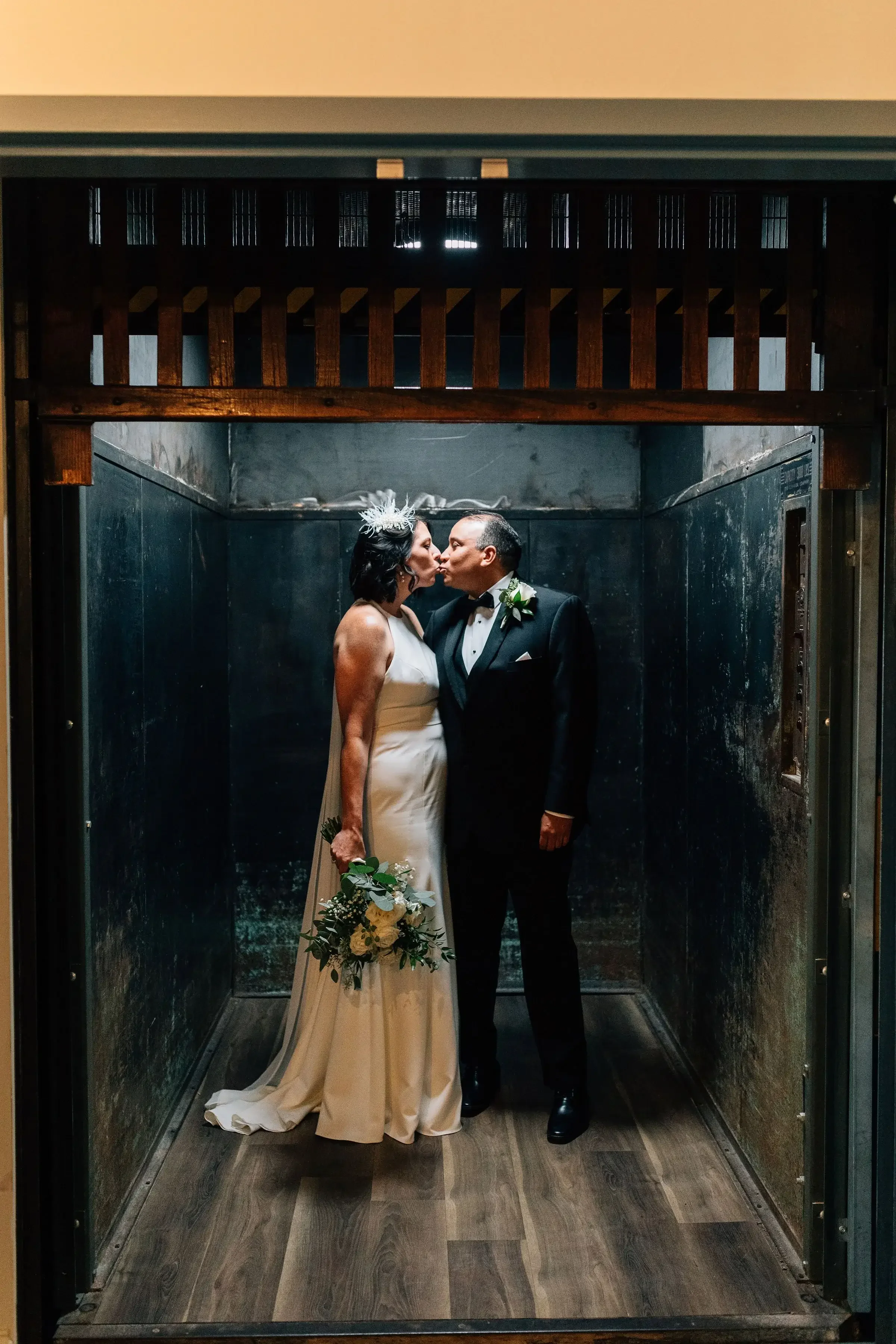 A bride and groom in wedding attire kissing inside an elevator, with the bride holding a bouquet of flowers.