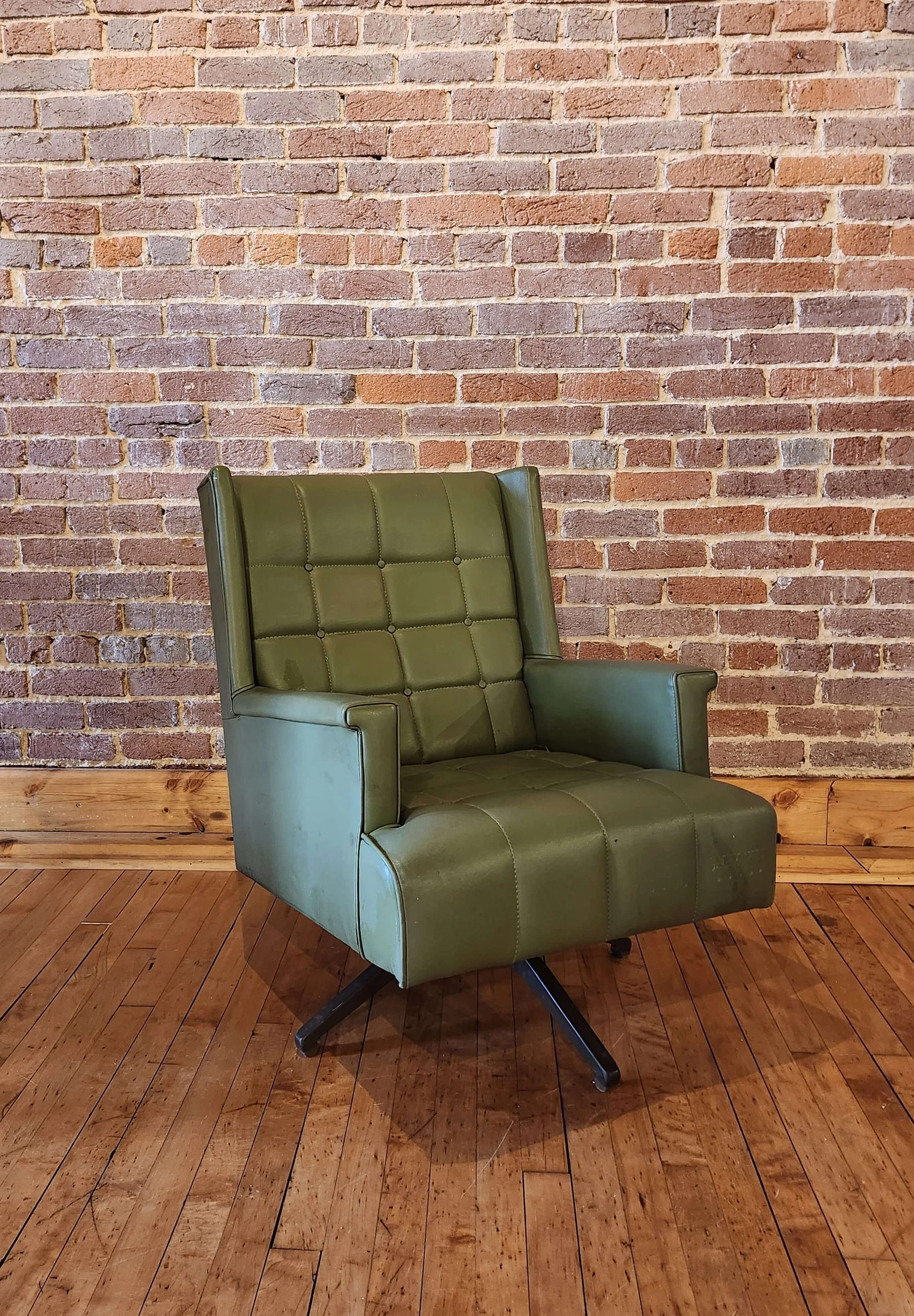 A green vintage-style office chair with a high back, armrests, and a swivel base, positioned in front of a brick wall and on a hardwood floor.