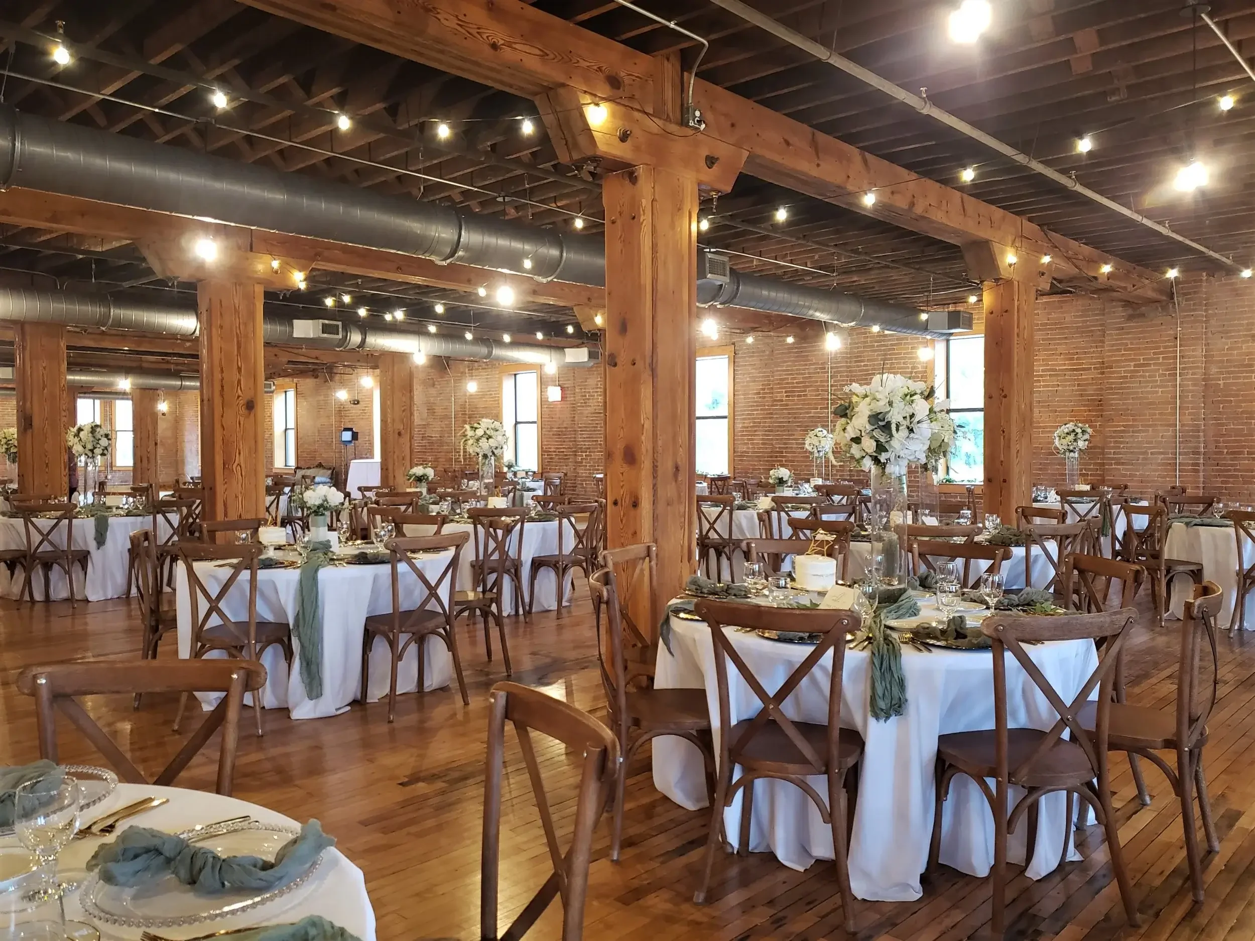 Event space with wooden beams, brick walls, large windows, and exposed ductwork, set up for a wedding or banquet with round tables covered in white tablecloths, decorated with tall floral centerpieces and place settings.