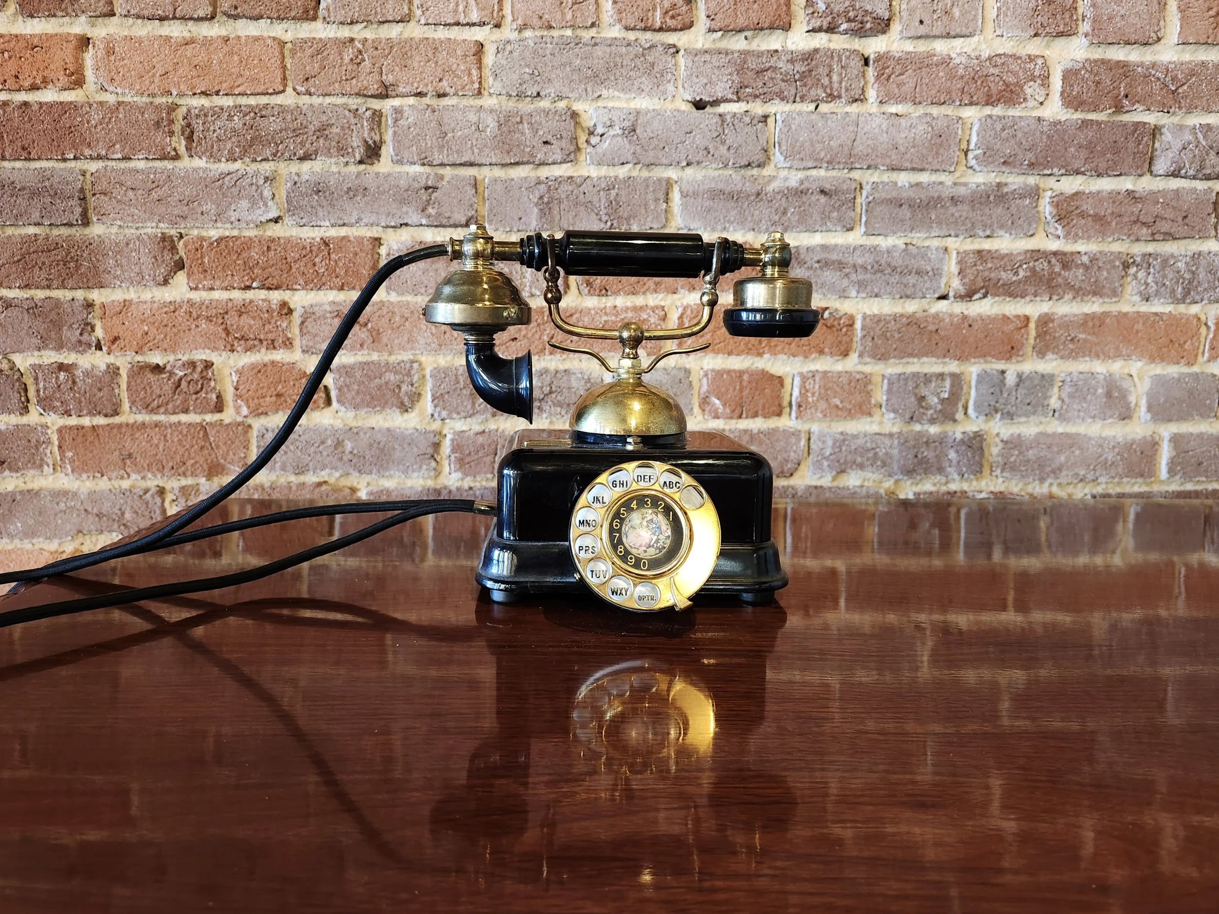 Vintage rotary telephone with black and gold accents on a wooden table, against a brick wall background.