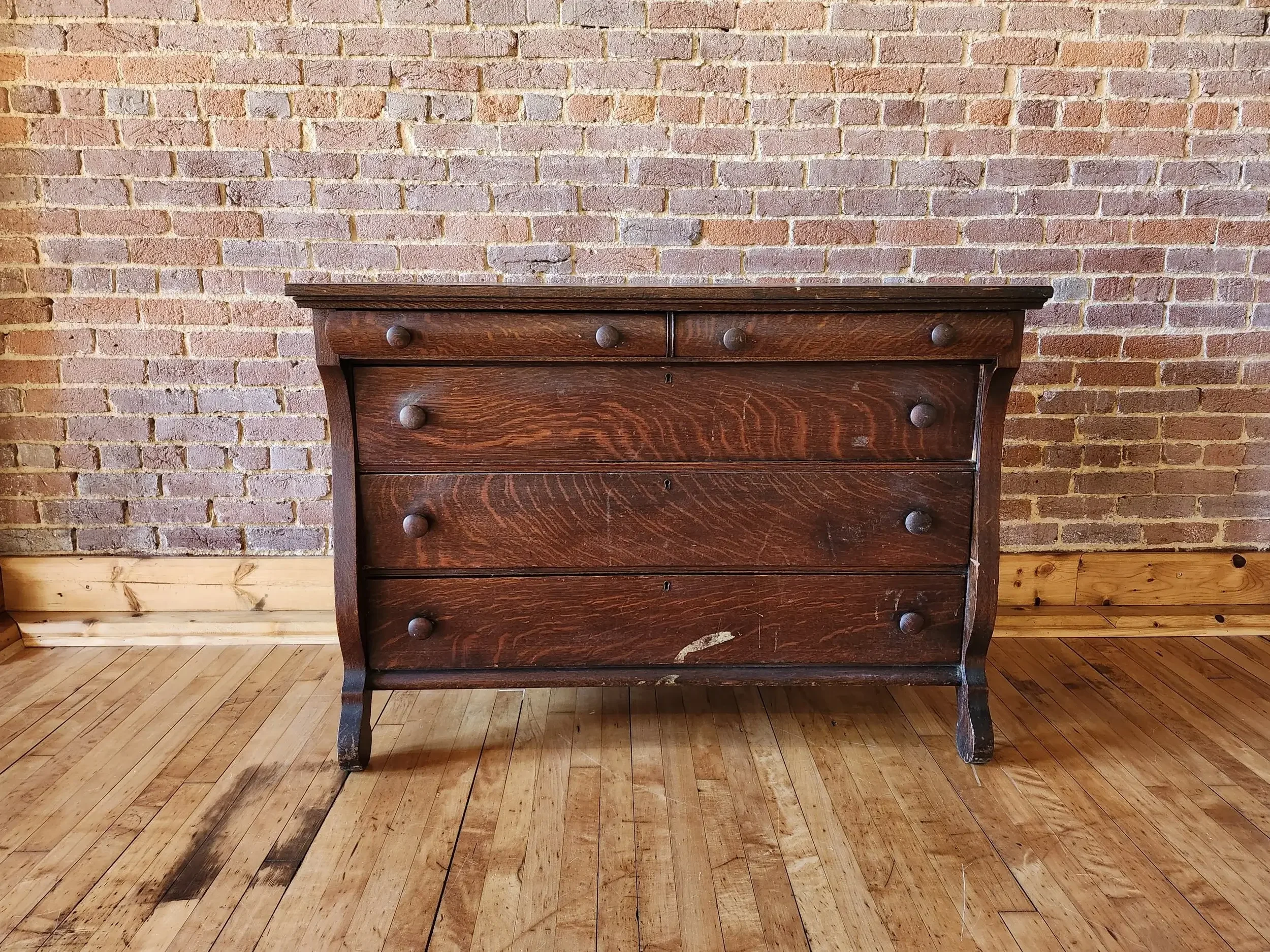An antique dark wood dresser with four drawers, positioned against a brick wall and standing on a wooden floor.