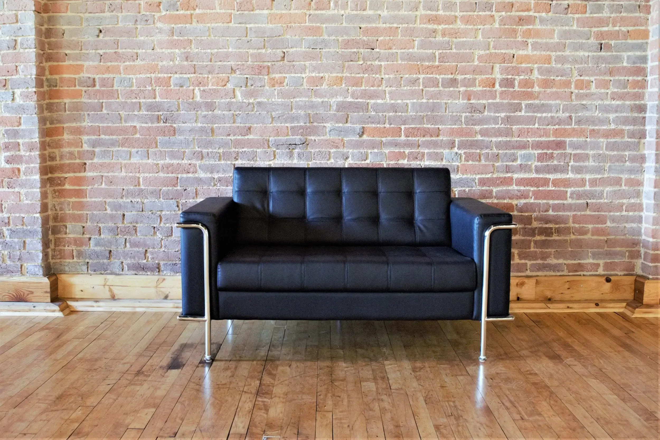 A black leather sofa with chrome armrests and legs, positioned against a brick wall with a wooden floor.