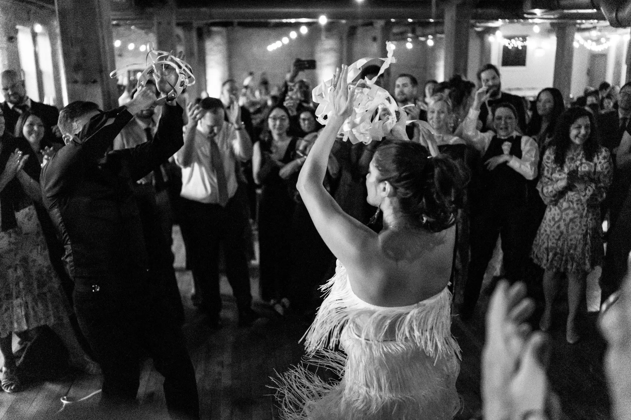 A woman in a fringed dress dances in front of a crowd at a party or celebration, holding streamers in her raised hand, while a man in a suit with a face mask dances nearby. The crowd watches and smiles in a lively indoor venue with string lights.