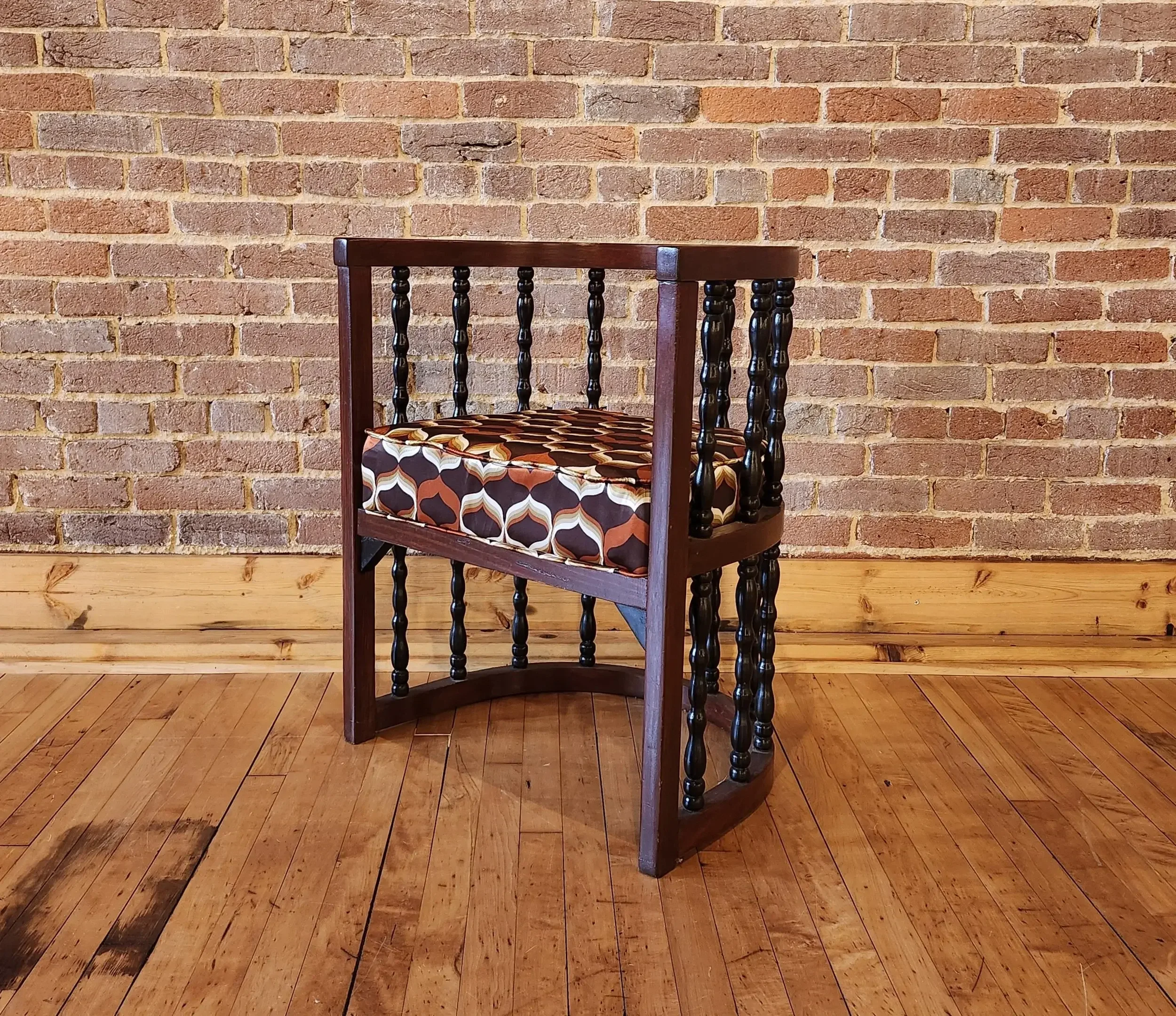 A wooden chair with black spindle legs and armrests, and a retro patterned cushion seat, placed on a hardwood floor against a brick wall background.