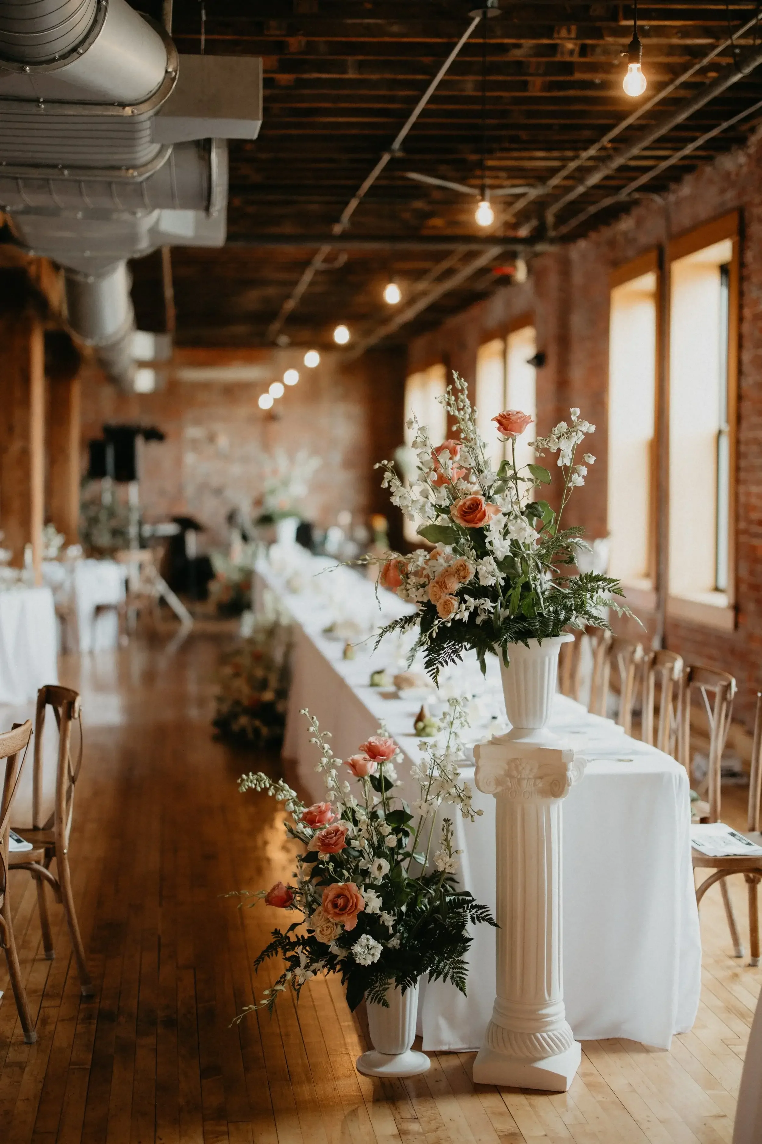 Elegant banquet hall with flower arrangements on tall white columns and a long head table, warm lighting, and exposed brick walls.