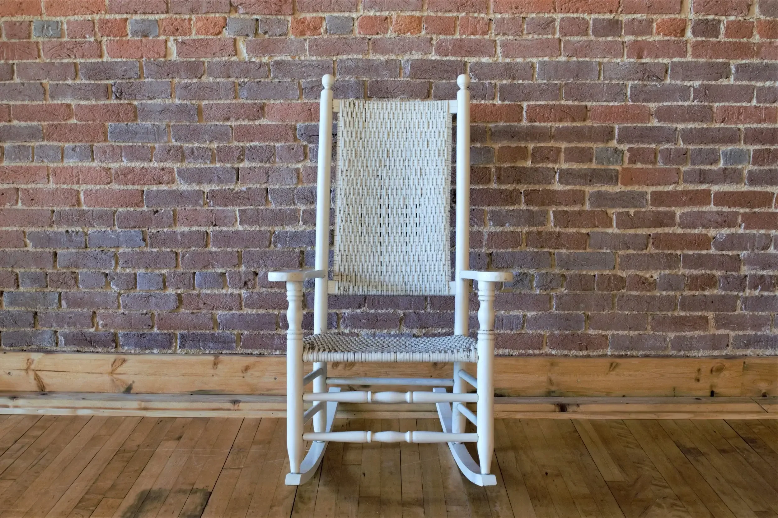 White wicker high-back rocking chair with armrests, positioned against a brick wall indoors on a wooden floor.