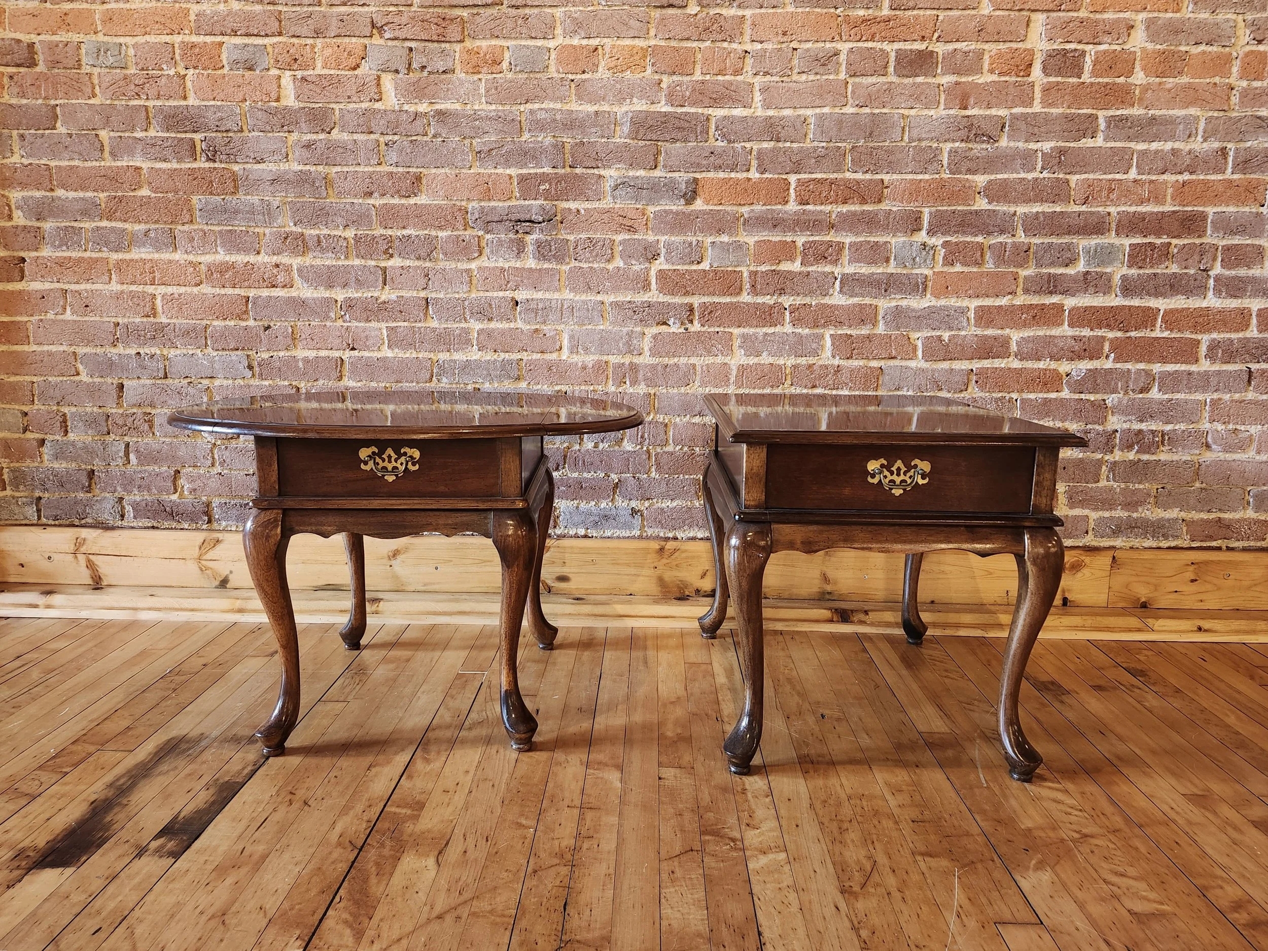 Two vintage wooden side tables with curved legs and brass handles, placed on a hardwood floor against a brick wall.