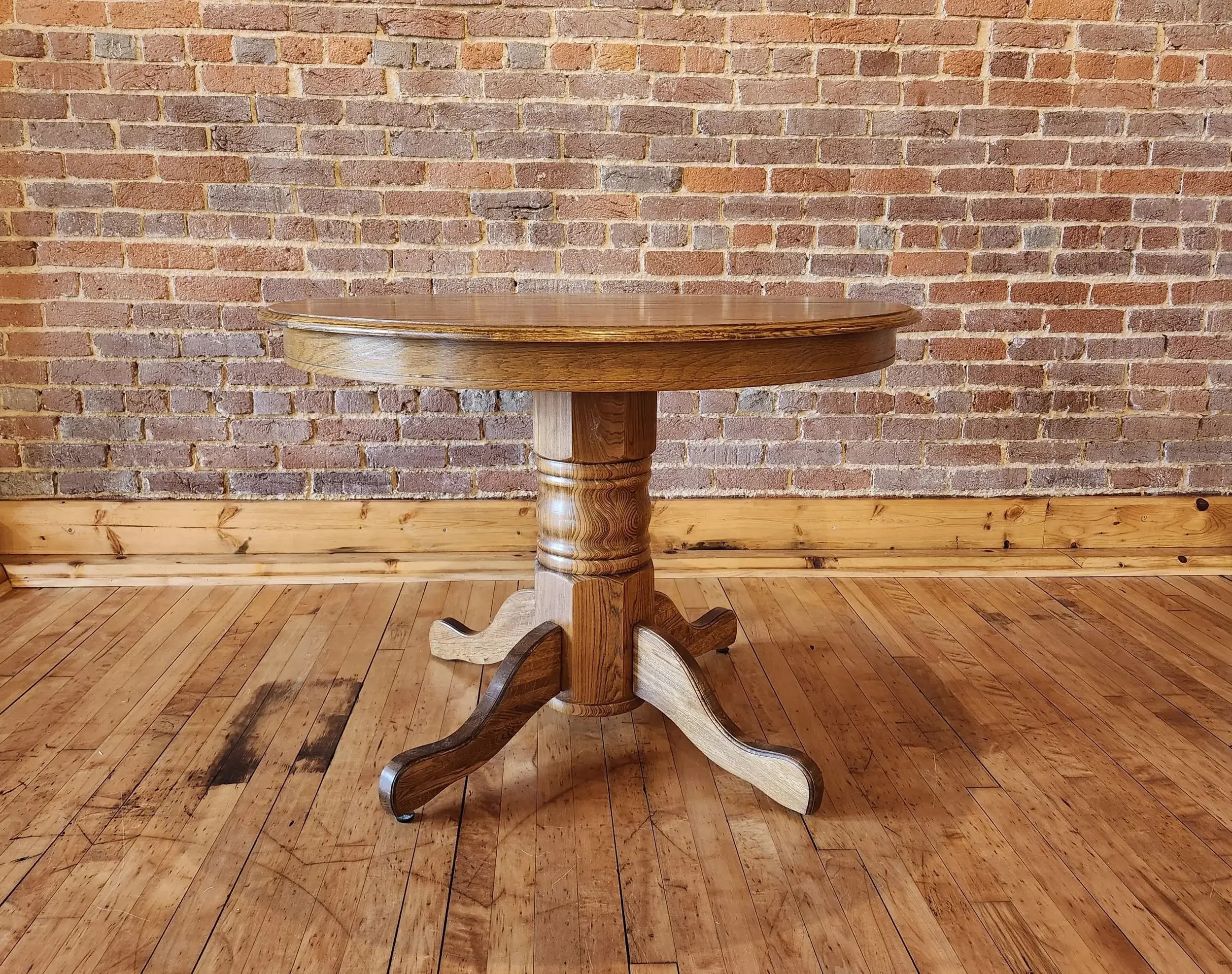 A round wooden table with a pedestal base and four curved legs, placed against a brick wall and wooden flooring.