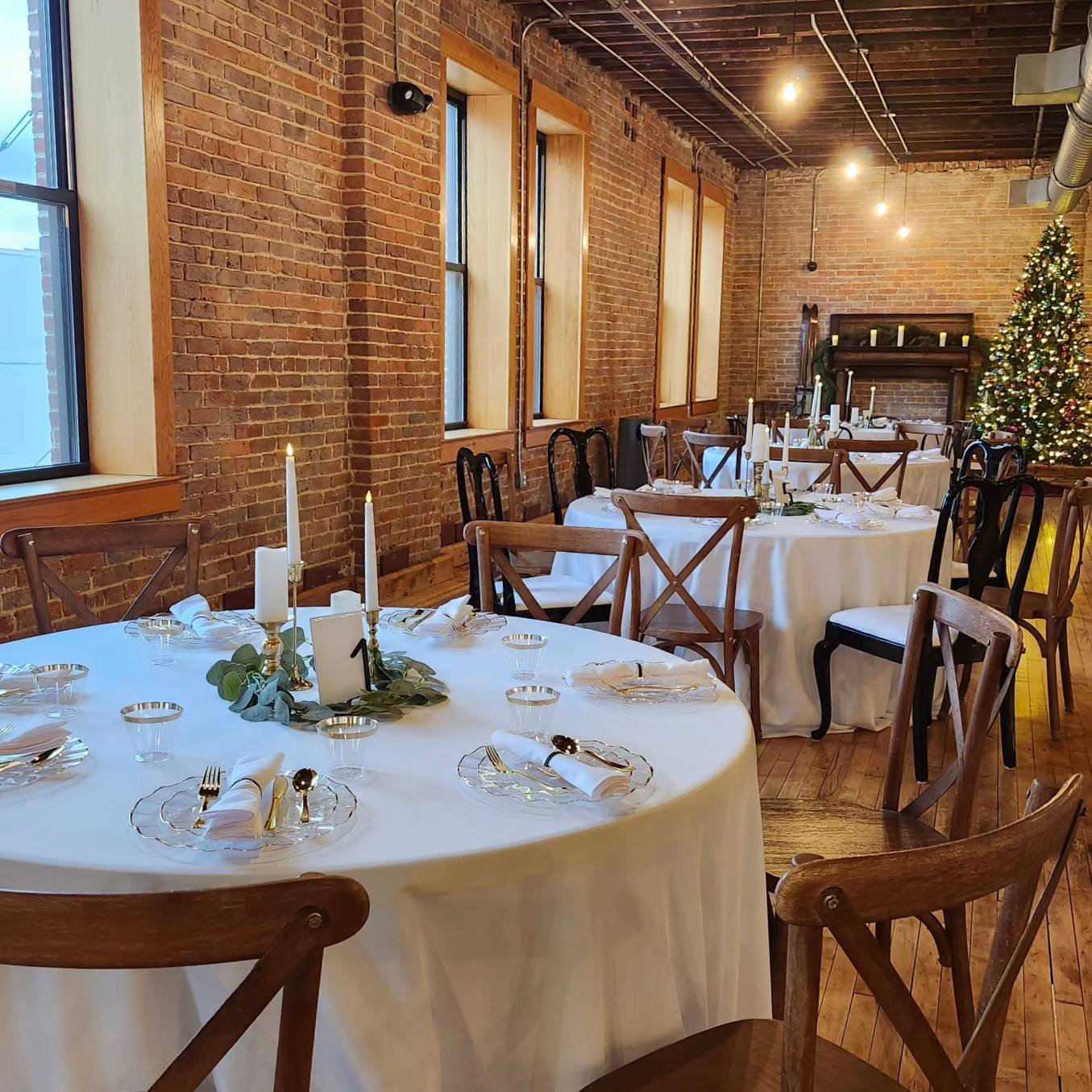 Decorated Christmas tree in a rustic banquet hall with round tables set for a holiday dinner.