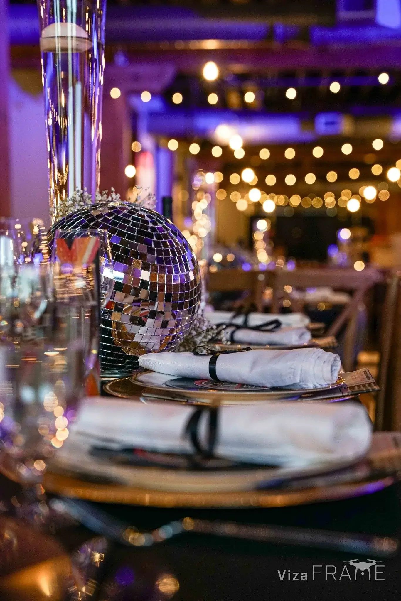 Elegant table setting at a party with white napkins, glassware, and a small disco ball centerpiece, with blurred string lights in the background creating a festive atmosphere.
