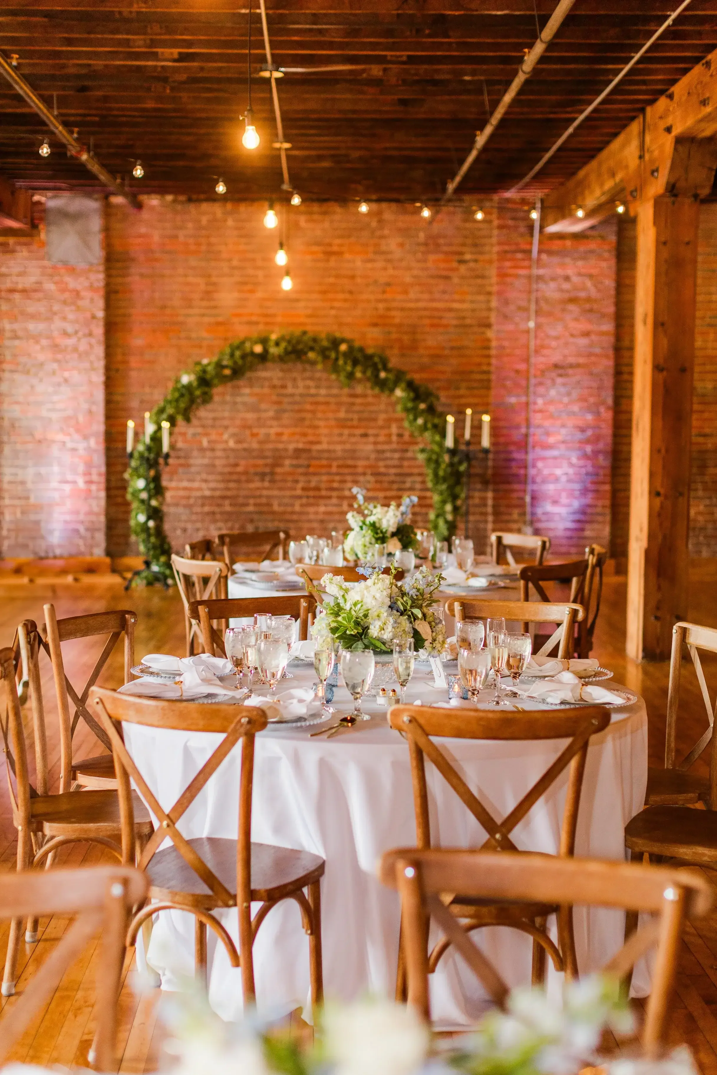 Round wedding reception table with white tablecloth, floral centerpiece, water glasses, and place settings, set in a rustic venue with brick walls, wooden beams, and decorative lighting.
