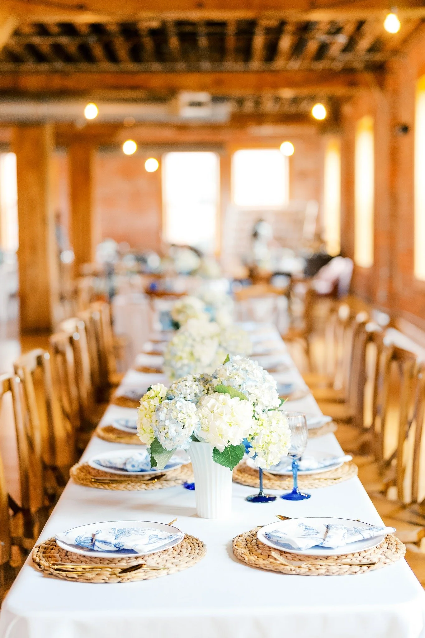 A long dining table set for a meal with a white tablecloth, floral centerpieces, plates, glasses, and gold utensils in a rustic wooden and brick room.