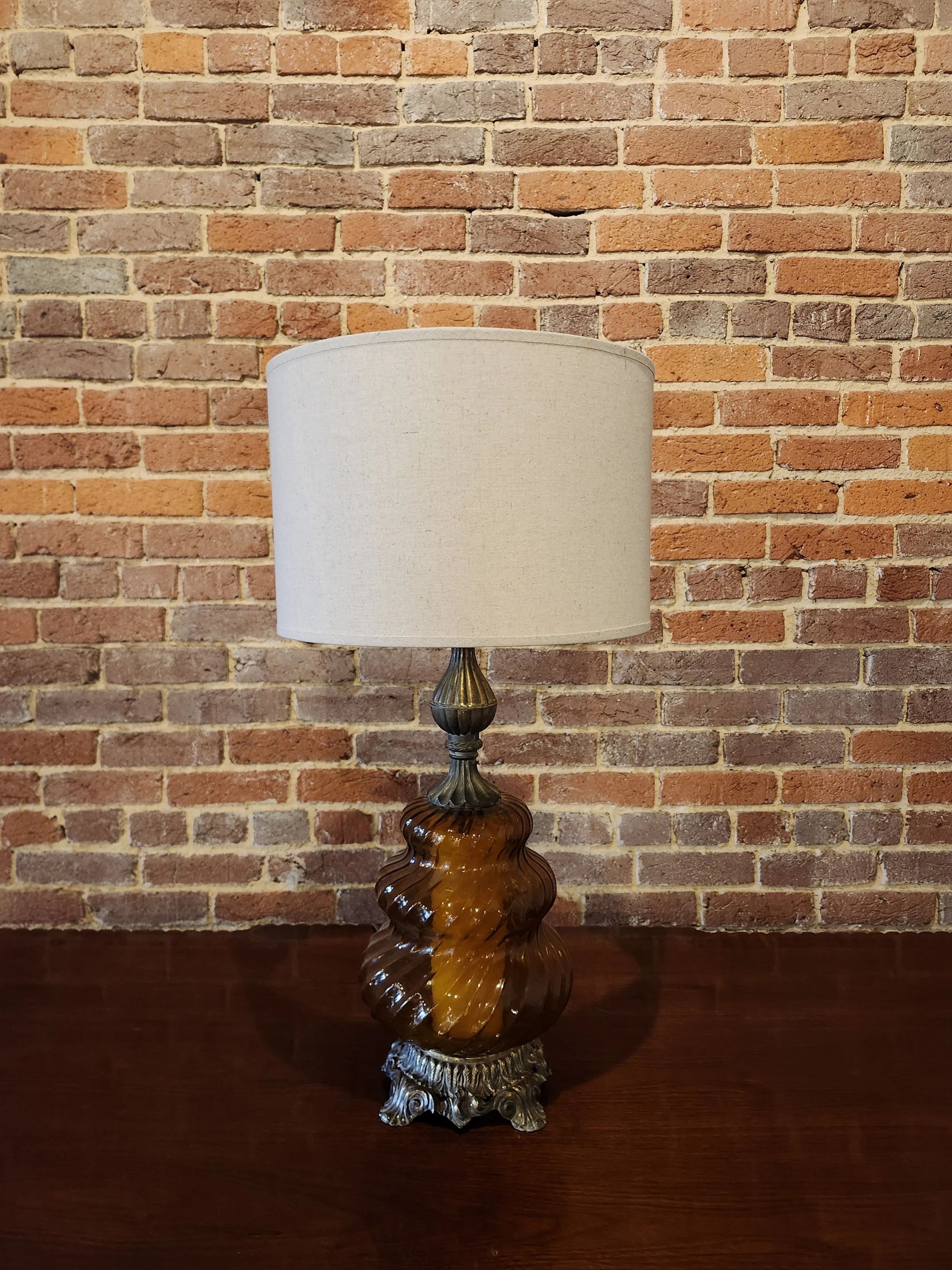 Vintage table lamp with a twisted amber glass base and a beige lampshade, placed on a wooden surface against an exposed brick wall.