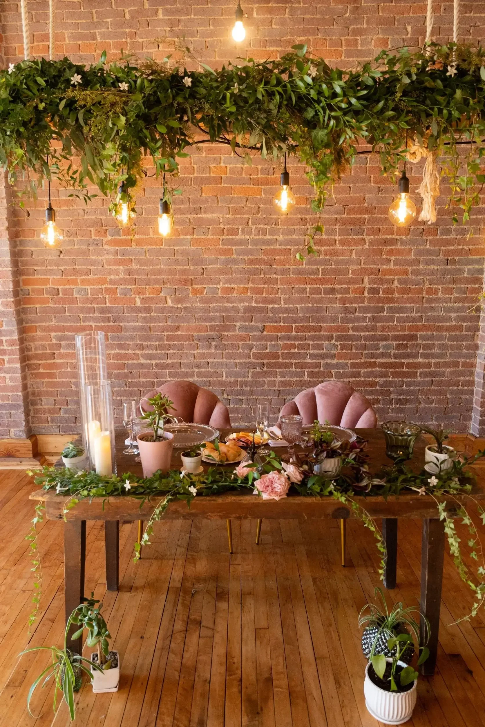 A rustic indoor dining table decorated with greenery, pink flowers, and potted plants against a brick wall. Hanging light bulbs above and chairs around the table suggest a cozy, elegant setting.