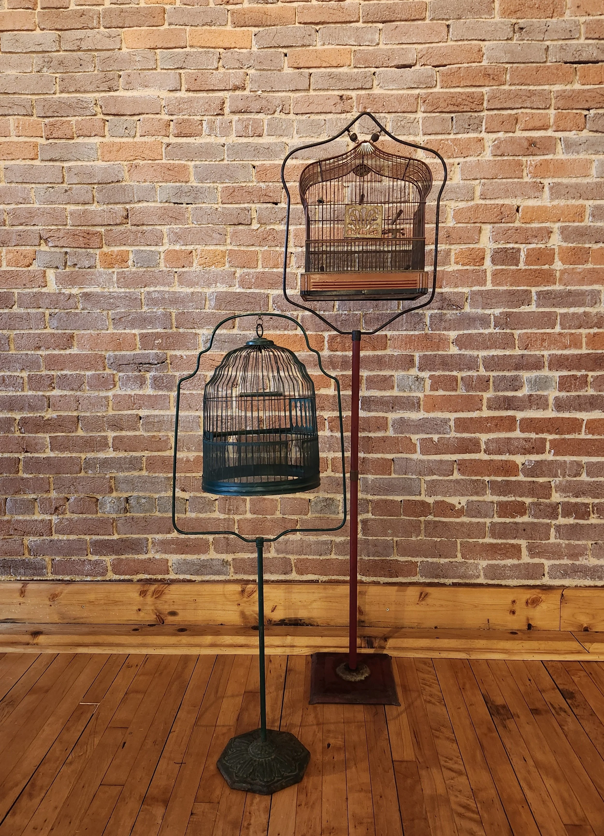 Two vintage birdcages on stands, one taller with a rounded top and one shorter with a flat top, placed against a brick wall in a room with wooden floors.