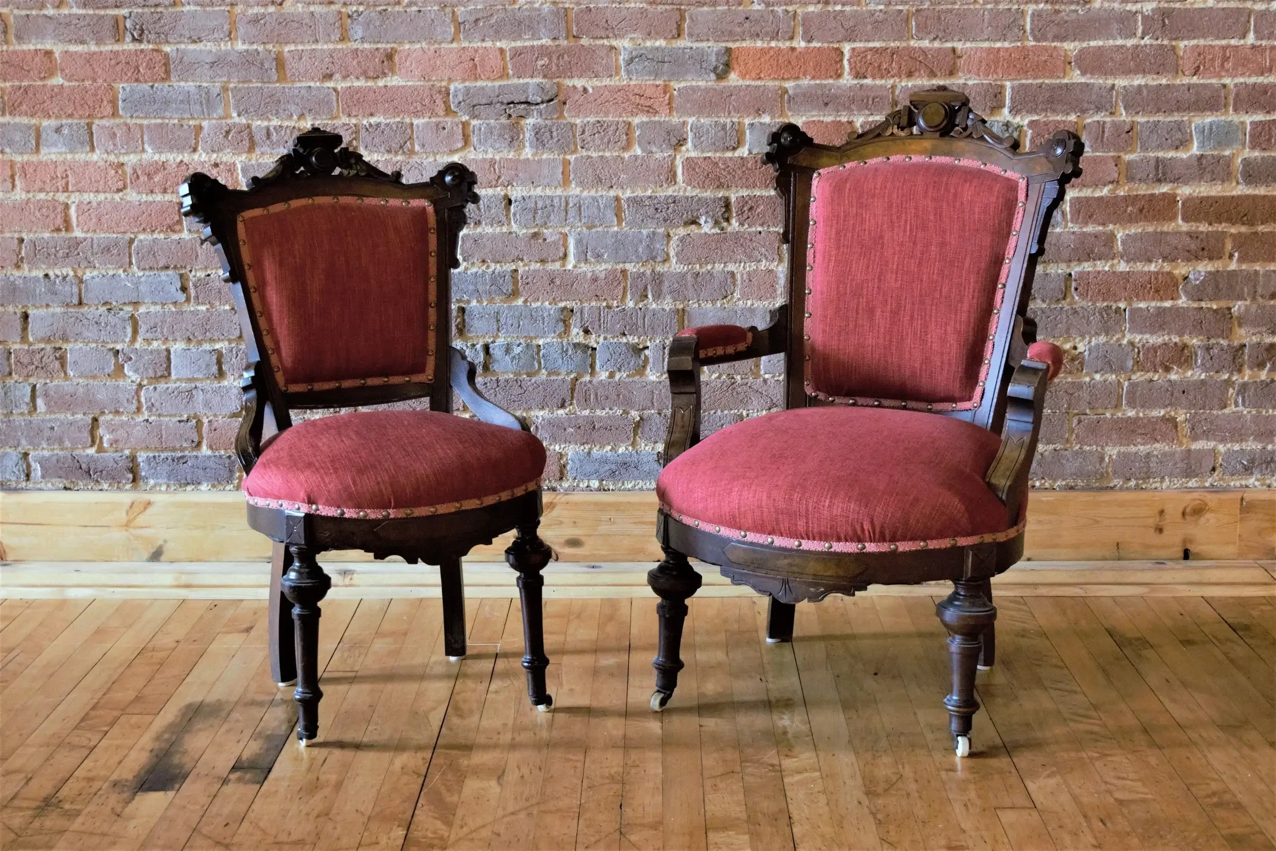 Two vintage wooden chairs with red upholstery set against a brick wall on a wooden floor.