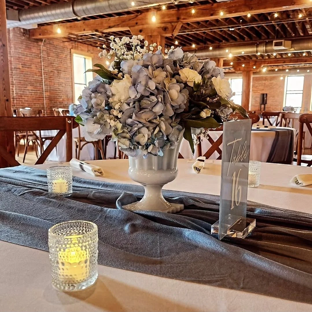 A wedding reception table decorated with a bouquet of white and light purple flowers in a white vase, surrounded by small candles in textured glass holders, with a table number sign reading 'Table 16' on a black table runner in a rustic venue with wo