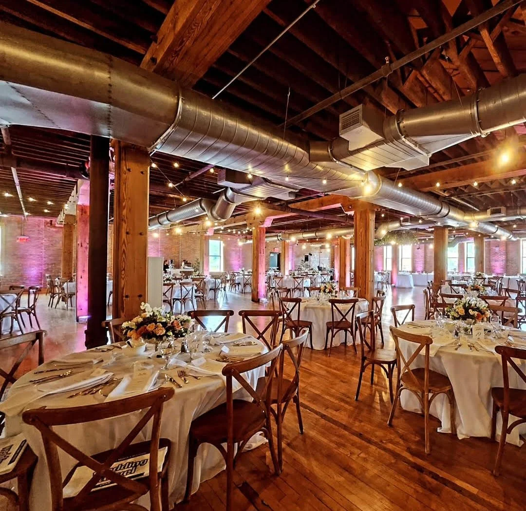 Wedding banquet hall with round tables, white tablecloths, floral centerpieces, and wooden chairs in a rustic, industrial-style venue with exposed ductwork, brick walls, and string lighting.