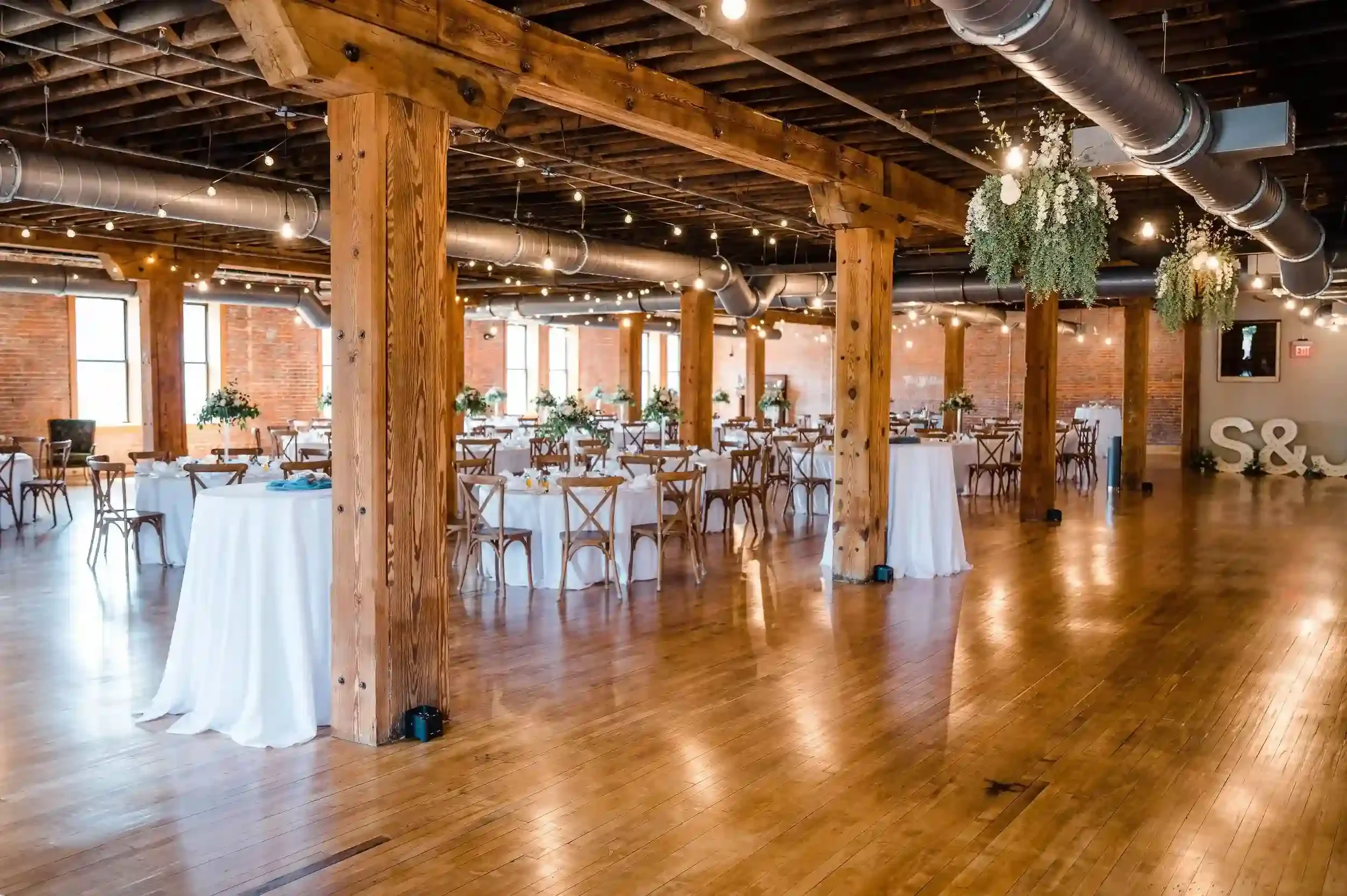 Event space with wooden beams, ductwork, string lights, hanging floral arrangements, and round tables with white tablecloths and chairs, set up for a celebration or wedding.