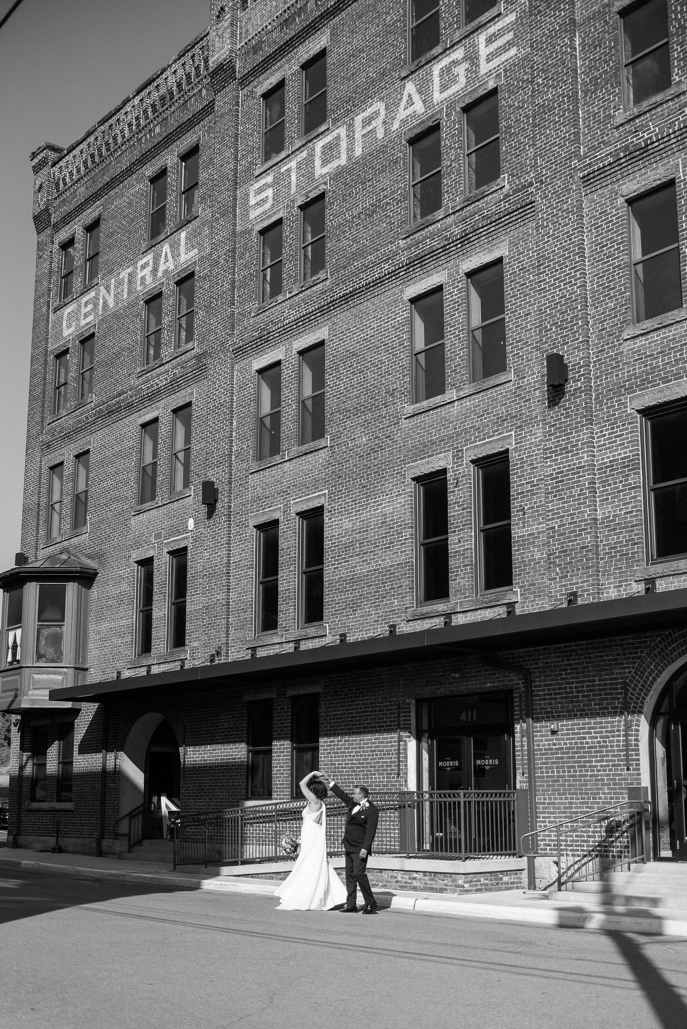 Black and white photo of a bride and groom standing outside a brick building with the sign "Central State Palace" visible on the upper wall.