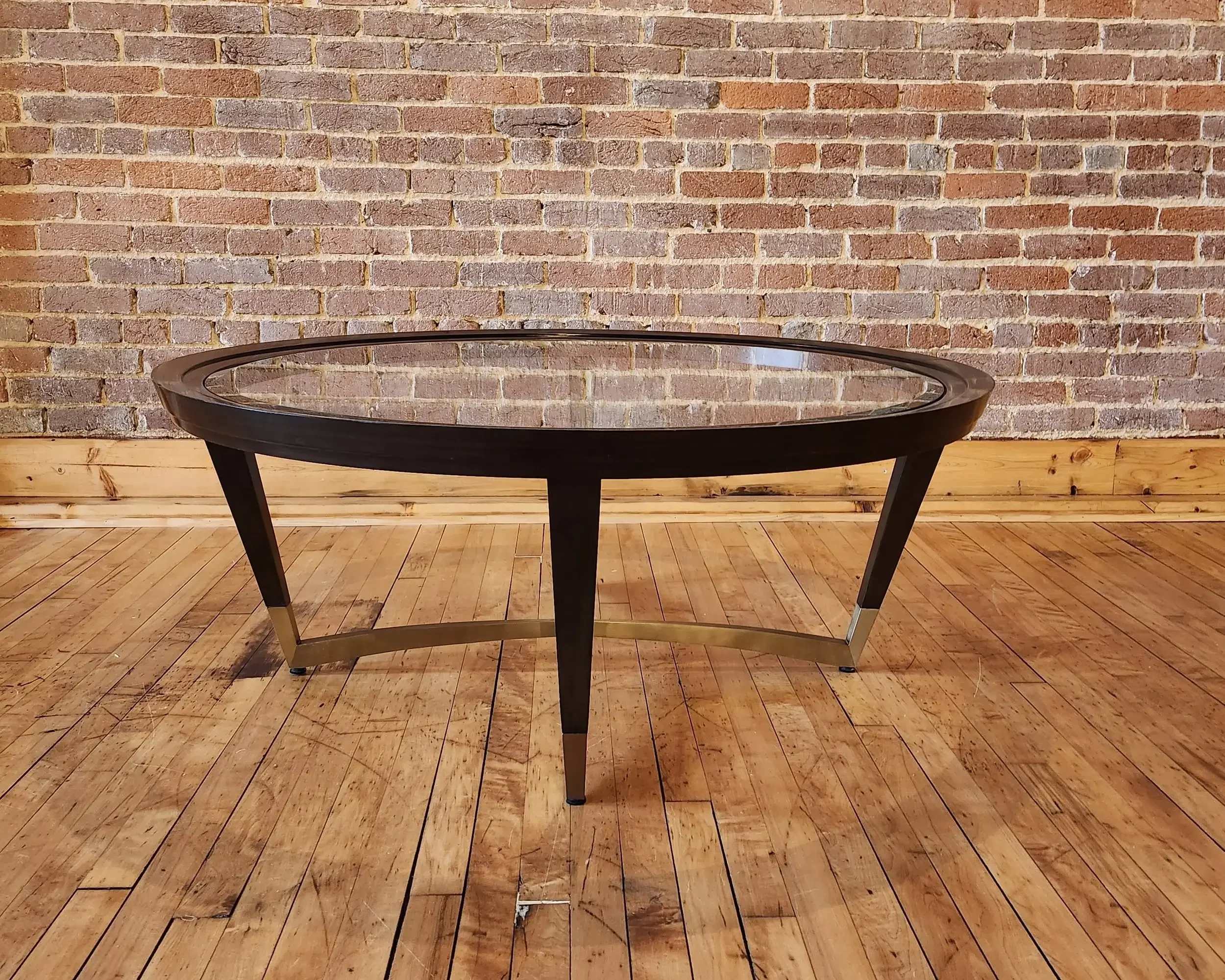 A modern oval-shaped glass coffee table with black and gold-metallic accents on the legs and edge, positioned on a wooden floor against a brick wall background.