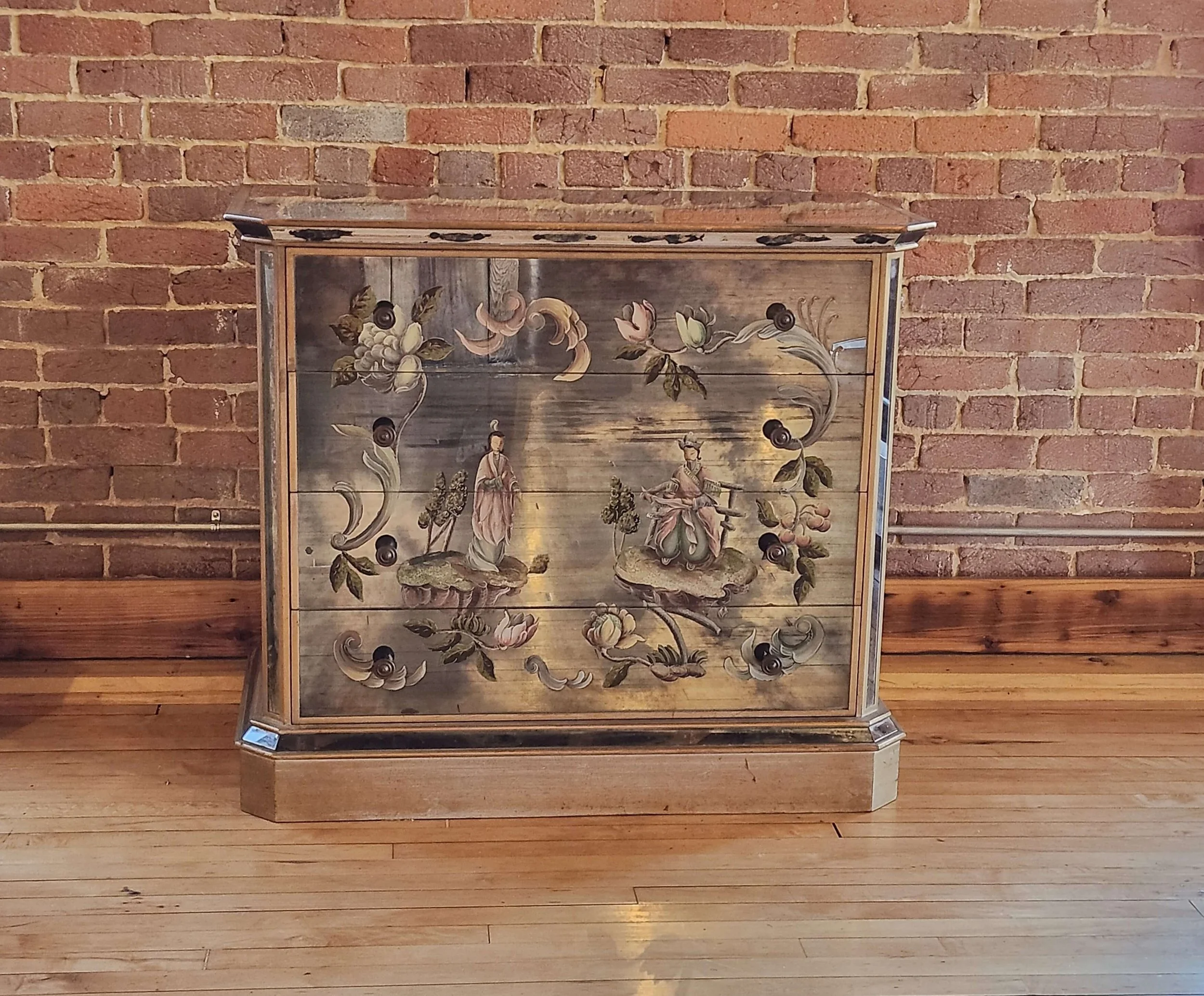 A wooden cabinet withhand-painted floral and figurative decoration, set against an exposed brick wall.