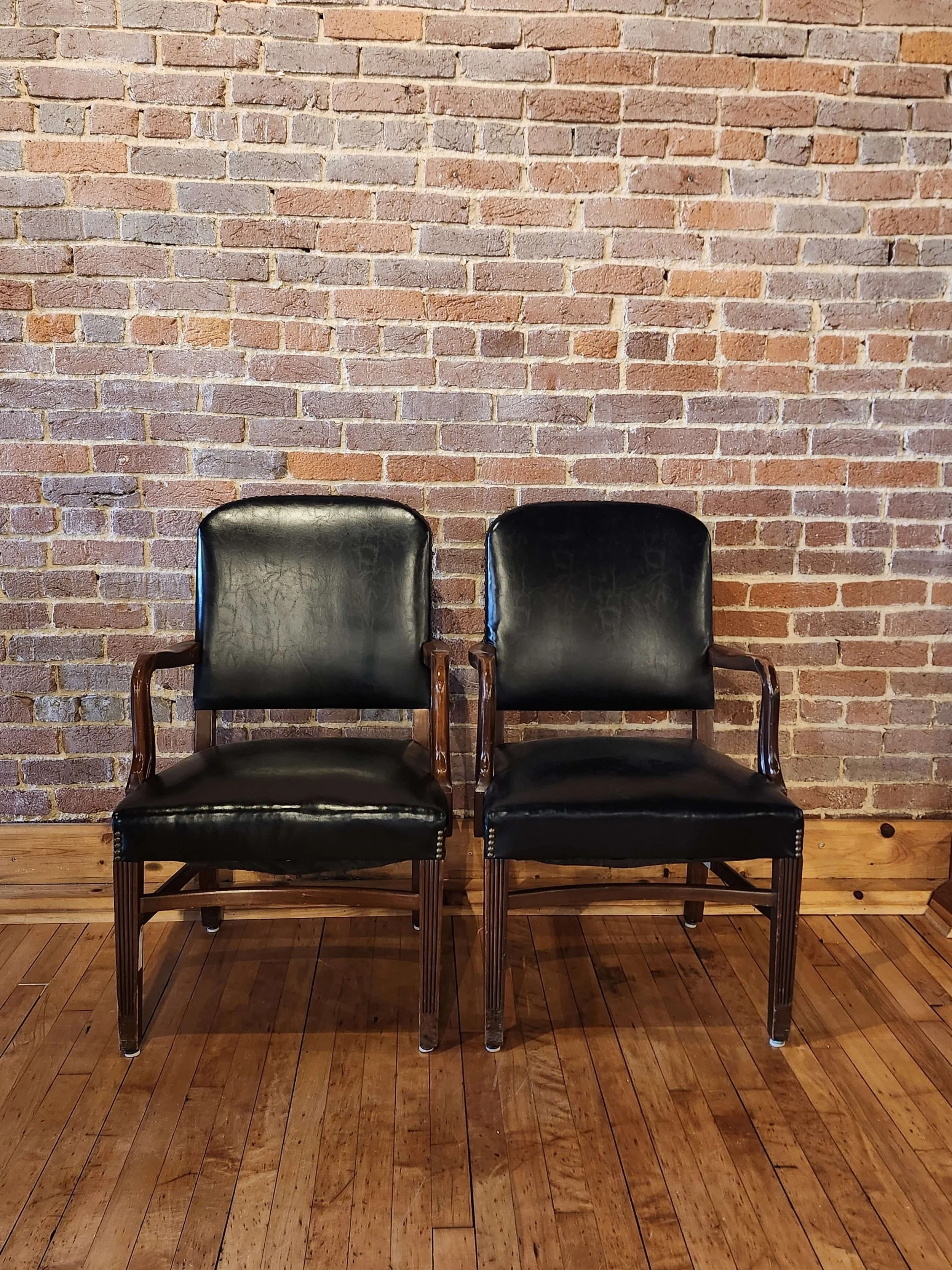 Two black leather chairs with wooden armrests and legs are placed side by side in front of a brick wall, on a wooden floor.