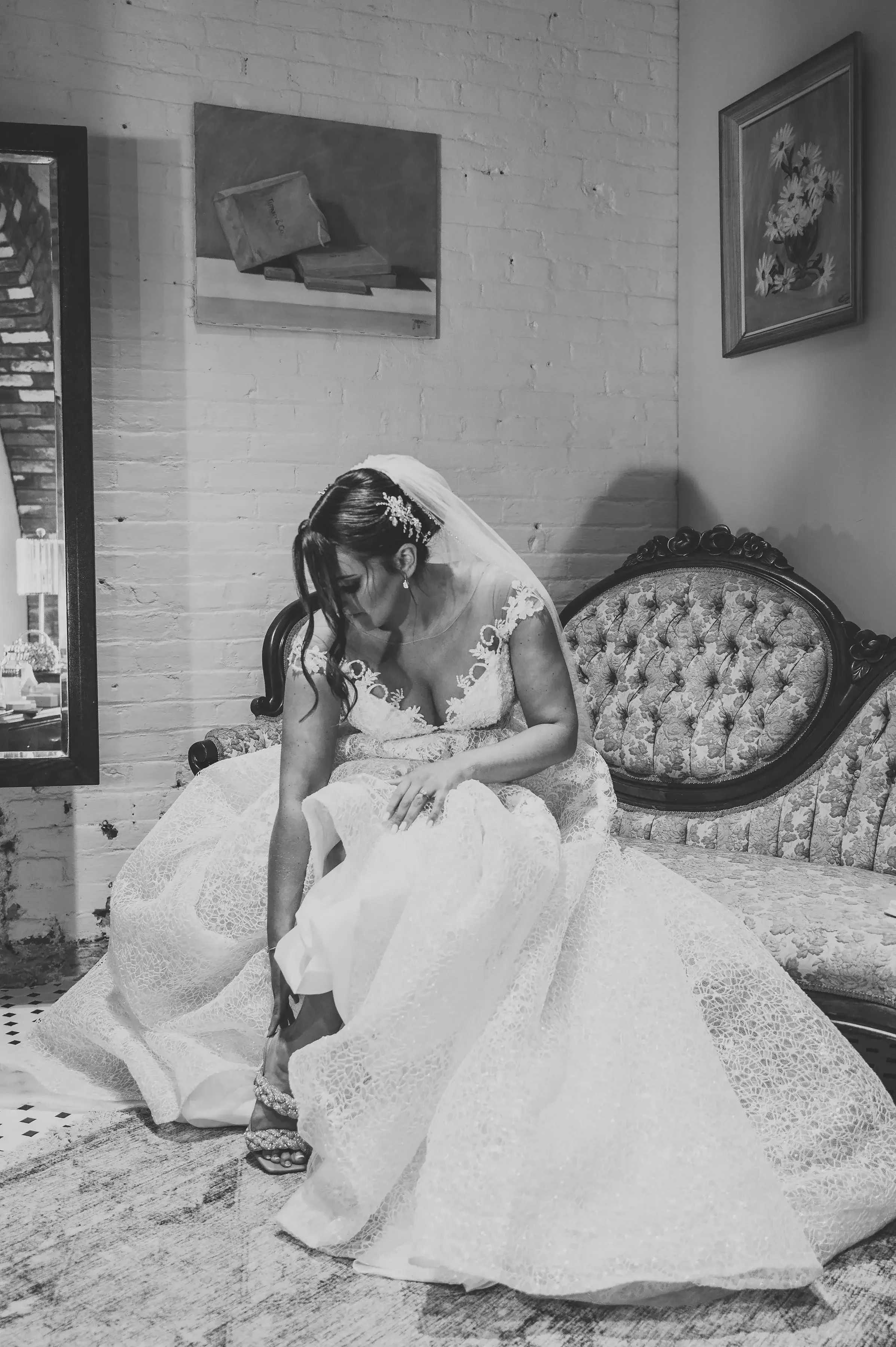 Black and white photo of a bride in a wedding dress sitting on an antique sofa, adjusting her shoe in a room with brick walls and framed artwork.