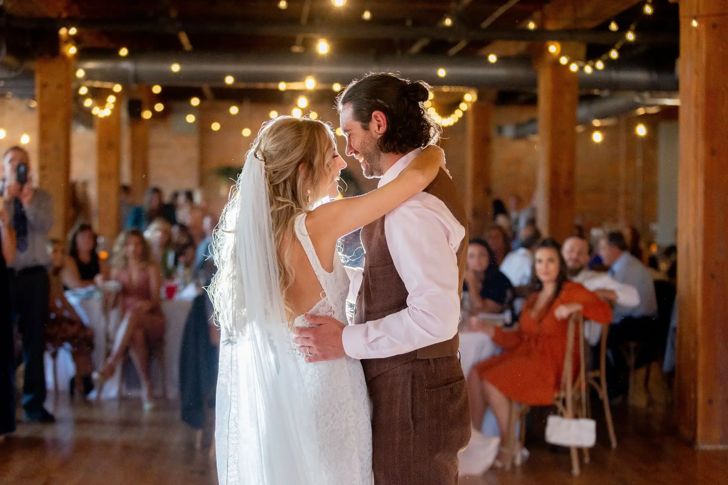 A bride and groom sharing their first dance at a wedding reception in a decorated venue with string lights and wooden beams, guests sitting at tables watching them.