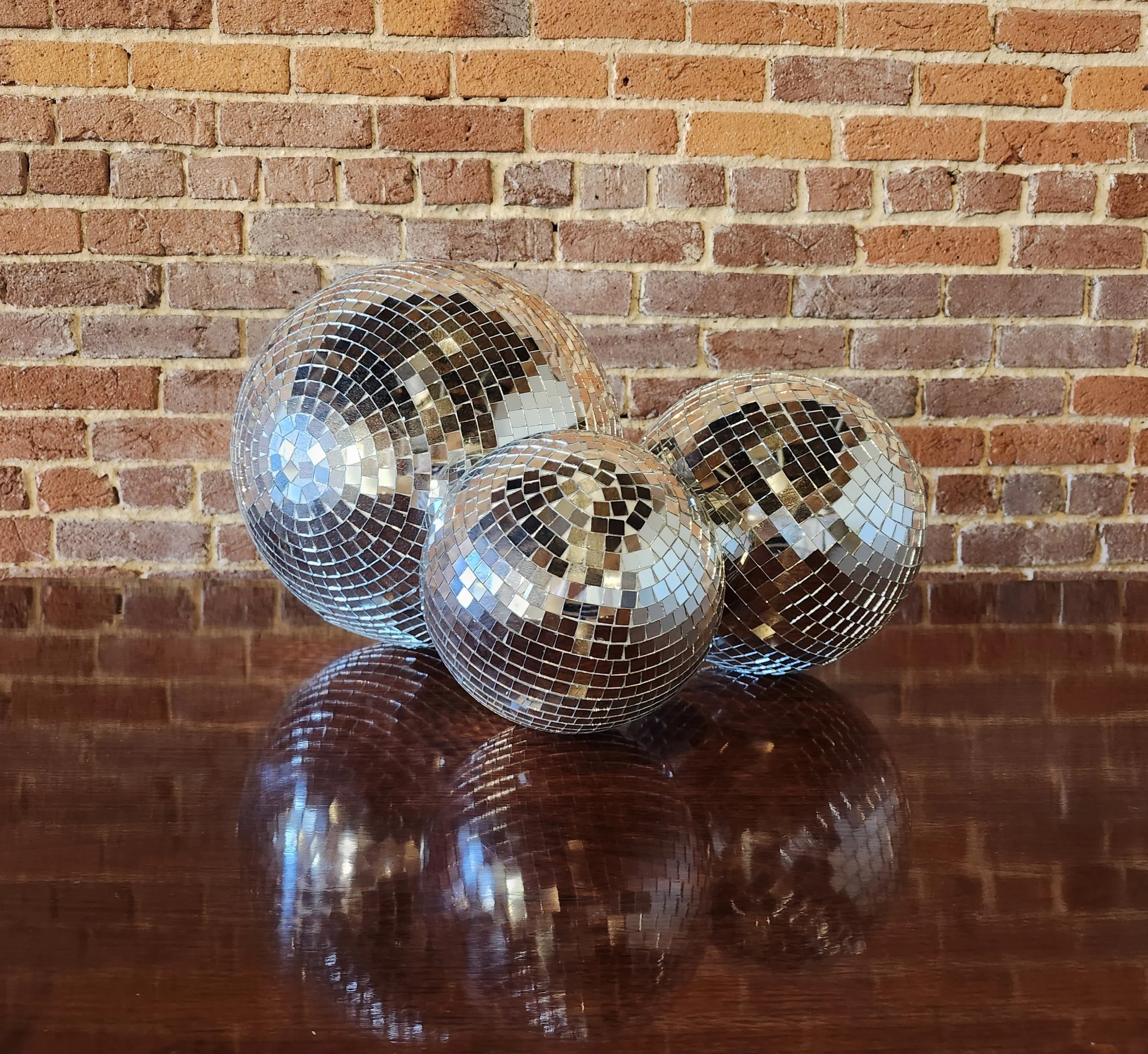 Four disco ball ornaments with mirrored mosaic tiles on a reflective wooden surface against a brick wall background.
