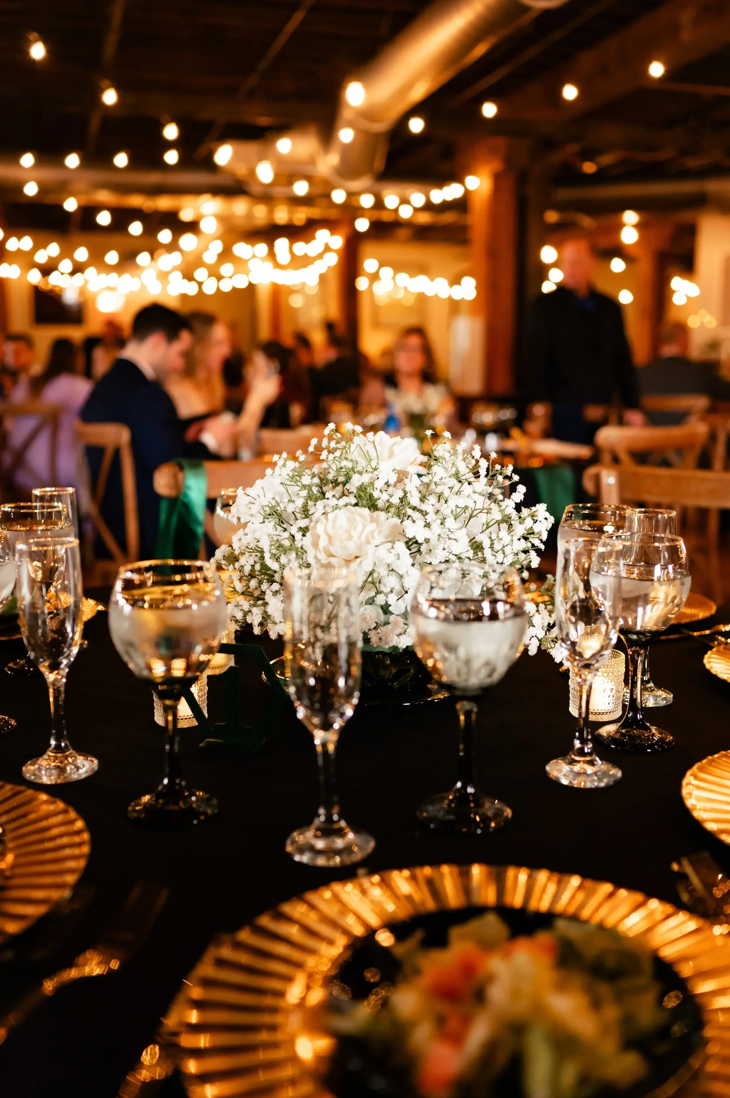 A wedding reception table decorated with a white floral centerpiece, multiple water glasses, and gold placemats, with blurred guests and string lights in the background.